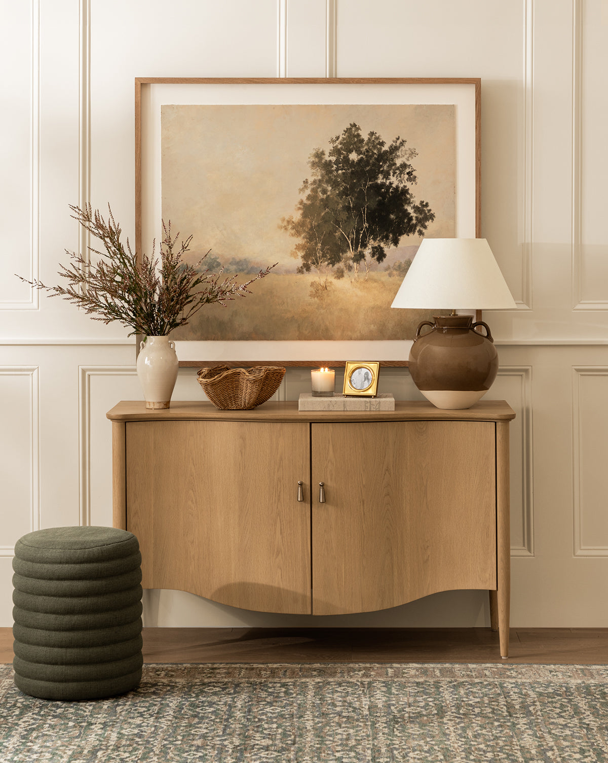A wooden sideboard topped with a lamp, vase of branches, basket, candle, clock, and landscape painting sits against a paneled wall. In front are a green pouf and the Made Goods Jefferson Stool on a patterned rug, all curated by Vendixs