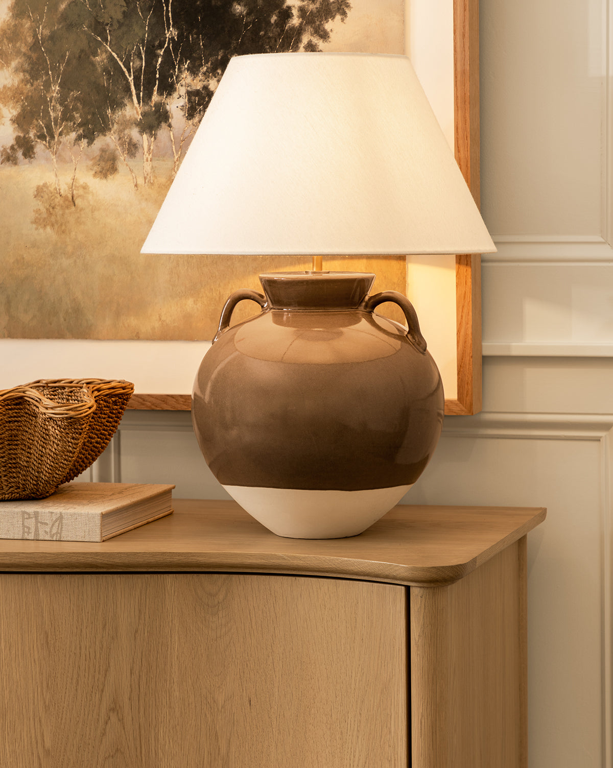 An Amphora Table Lamp in brown and beige ceramic with a white shade is displayed on a light wood cabinet beside a woven basket and book, with a landscape painting hanging on the wall behind, by Vendixs