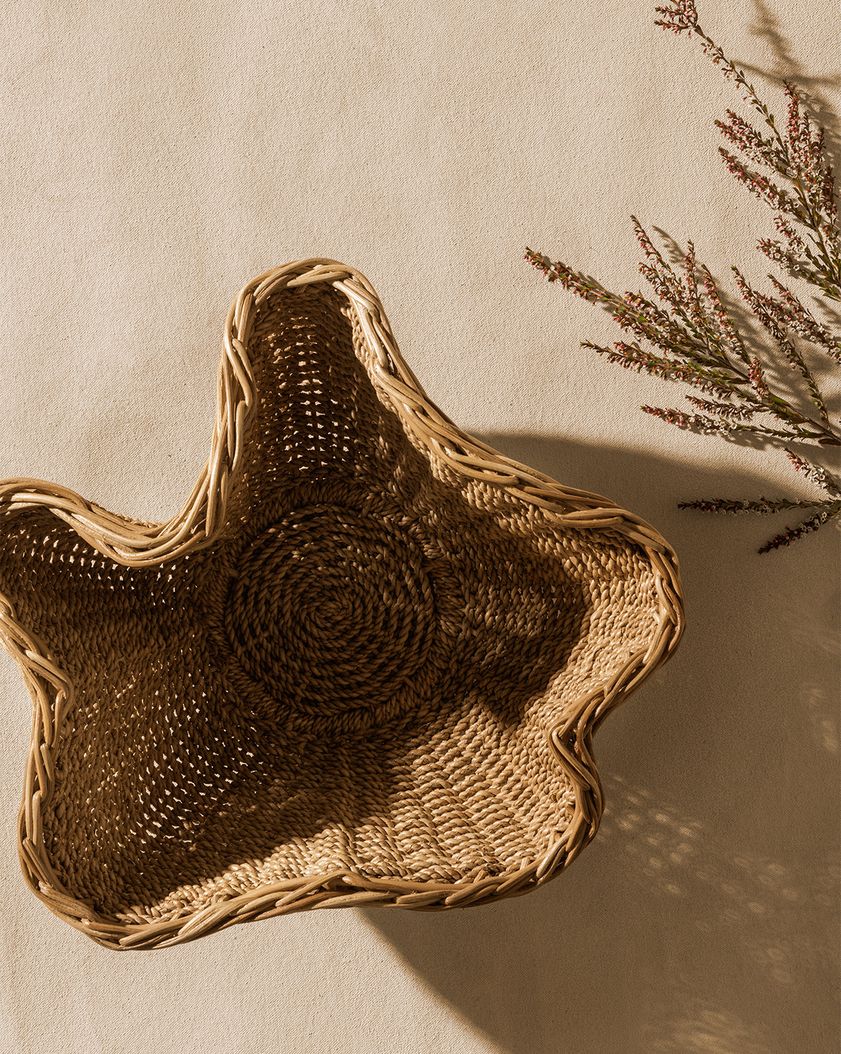 The Goulding Woven Bowl, shaped like a star, rests on a beige surface beside delicate flowers casting soft shadows—ideal as charming entryway decor, by Vendixs