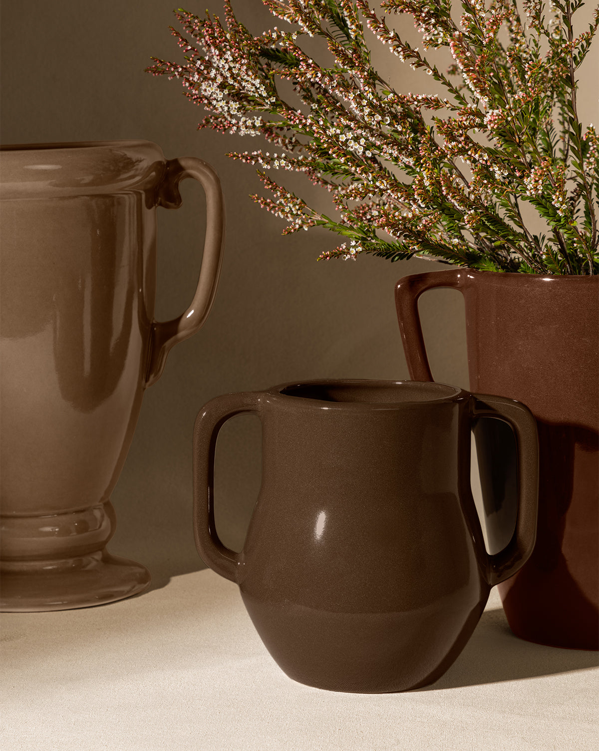 The Victoire Vase, an elegant earthenware vase in earthy brown tones, is displayed with two ceramic mugs on a neutral surface. One mug contains green foliage with white and pink flowers, creating a warm, cozy atmosphere under soft lighting, by Vendixs