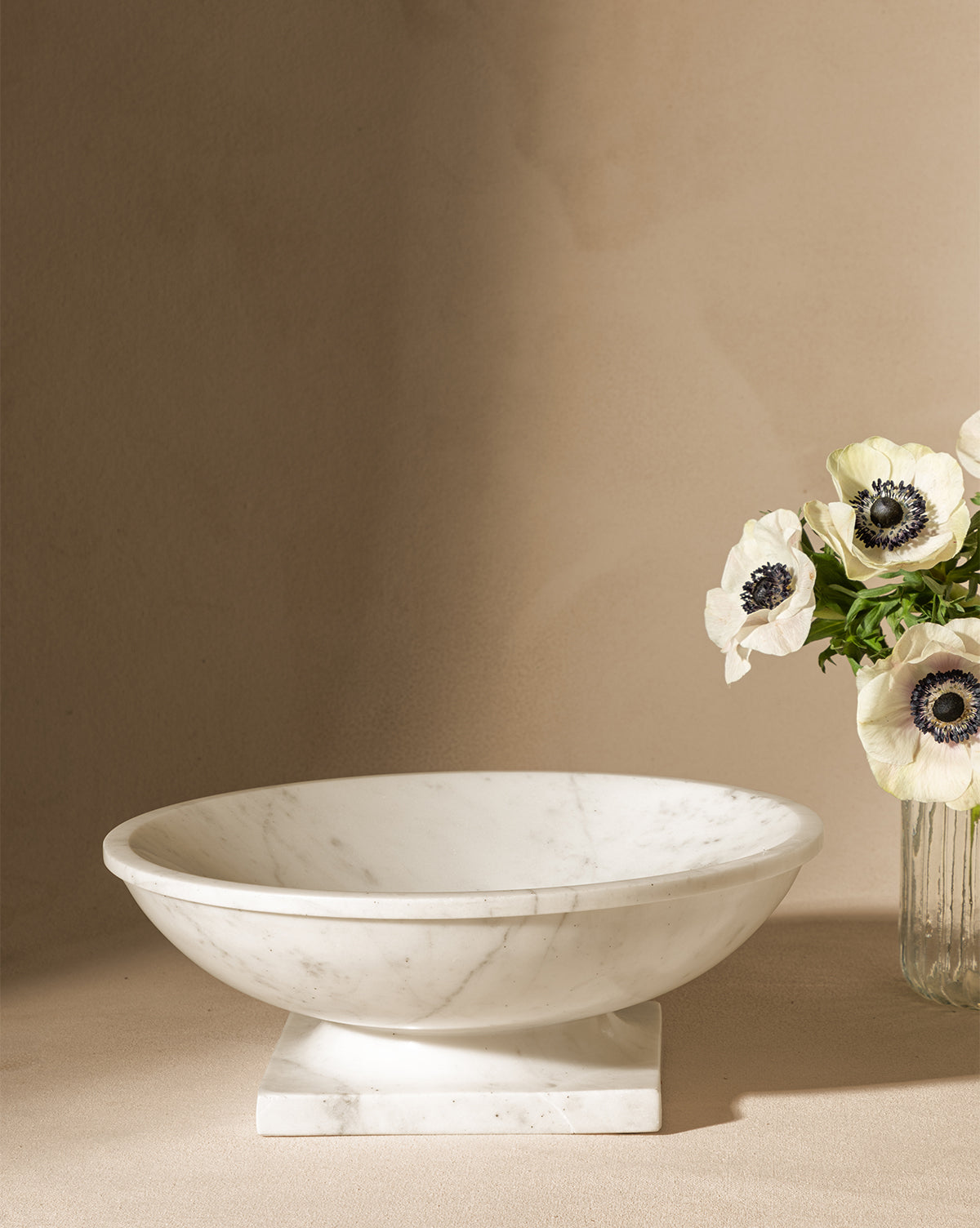 The Tayva Bowl, a sculptural white marble piece with a square base, rests on a neutral surface beside a glass vase of white anemones with dark centers against a beige background, by Vendixs