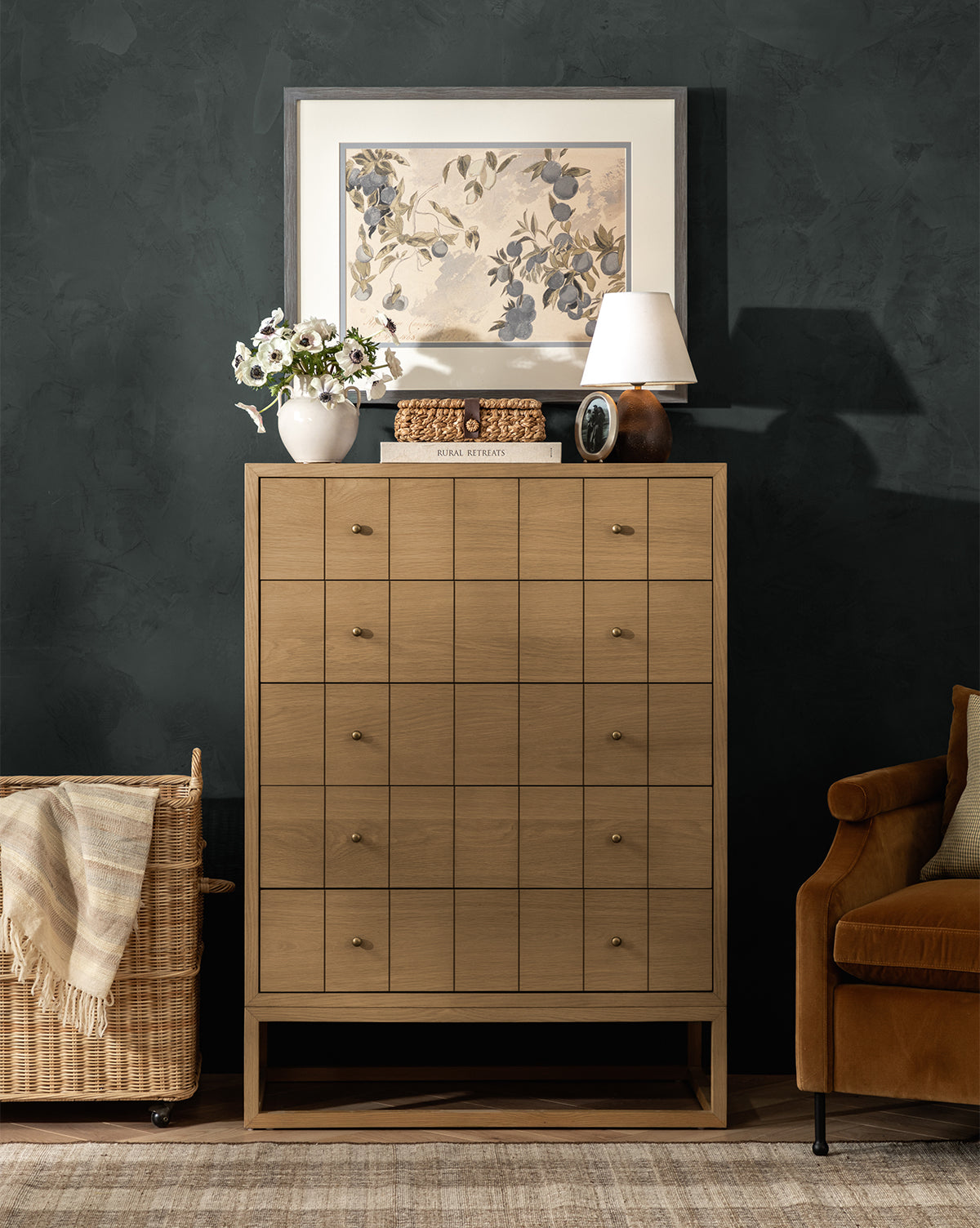 A light wood chest with twelve drawers stands against a dark wall. On top are a lamp, books, a white vase with flowers, decor, and Blue Fruits. Above hangs botanical art in a charcoal woodgrain frame. A wicker basket and brown armchair are nearby, by Vendixs