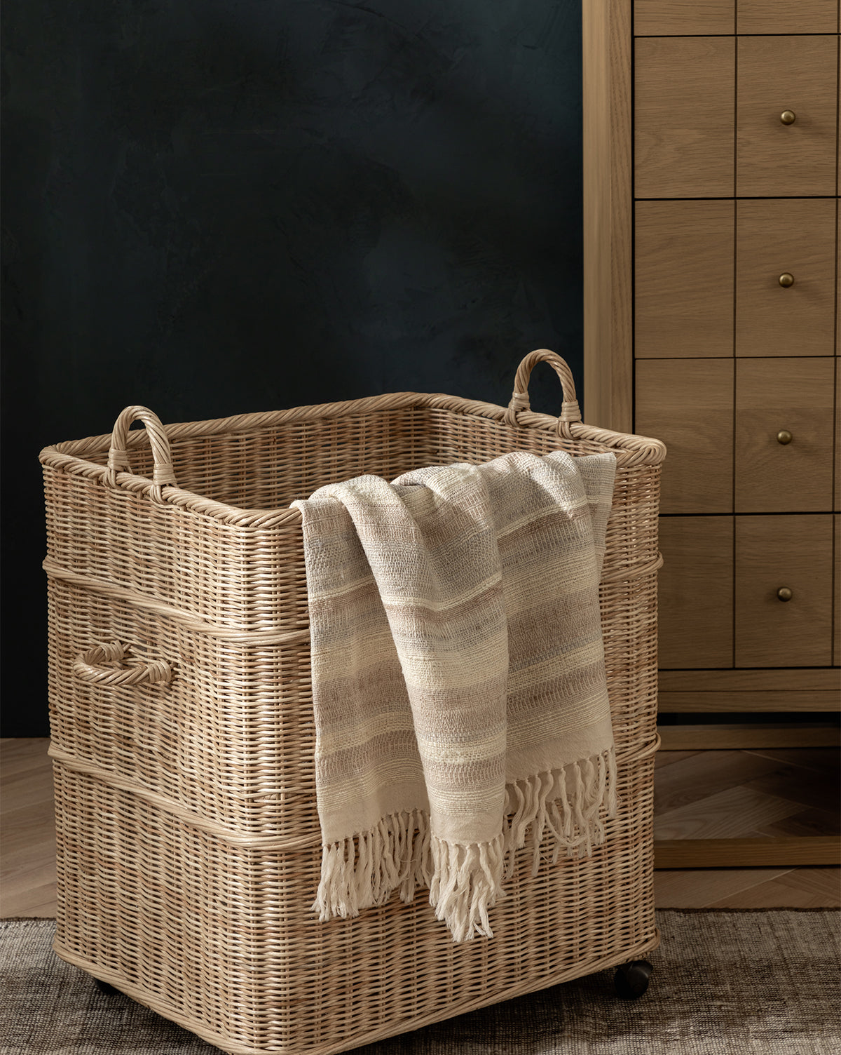 The Chiara Laundry Basket with natural wicker finish, handles, and wheels sits on a wooden floor. A cream fringed blanket hangs over it, while a wooden dresser with brass knobs completes this cozy laundry room scene, by Vendixs