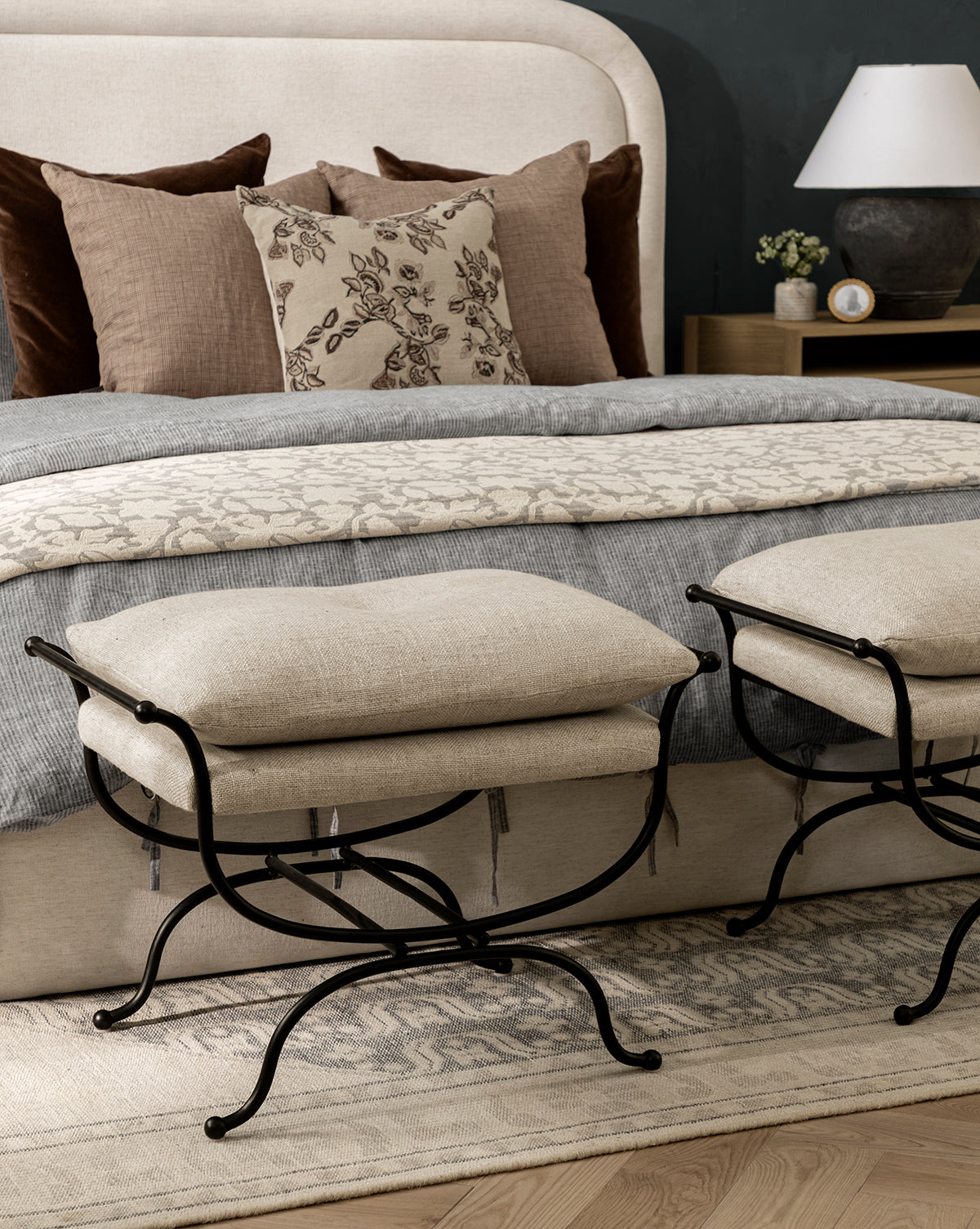 A cozy bedroom features a beige upholstered bed, layered brown and patterned pillows, a gray blanket, two Makerspalm Alameda Stools with matte black iron bases, plus a nightstand with a lamp and a decorative rug on the wooden floor, all curated in the inviting style of Vendixs