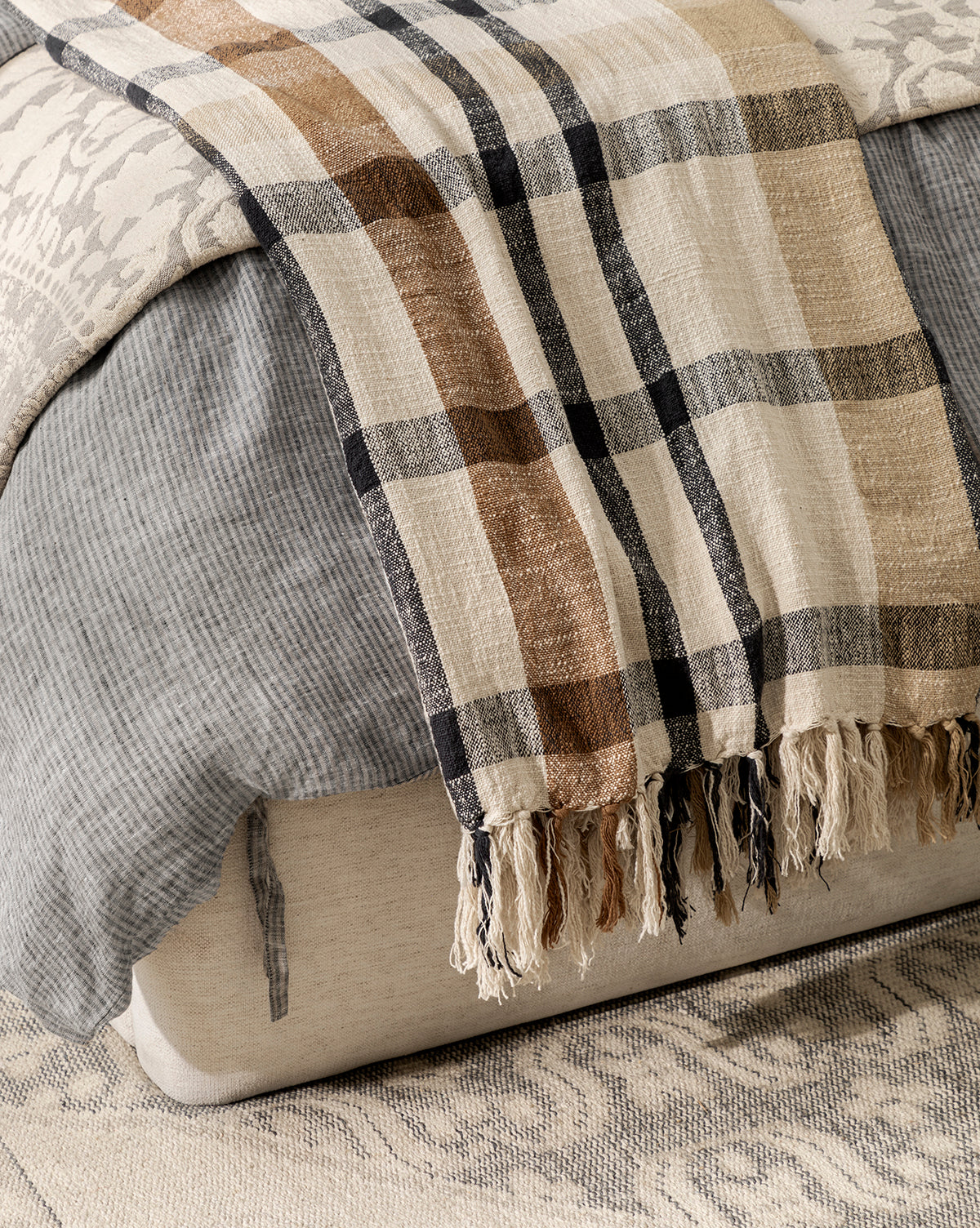 A close-up of a bed features layered bedding, including the Raj Albright Plaid Throw—a cozy cotton accent with brown and black stripes, fringed edges, and draped over the foot for added texture. Vendixs