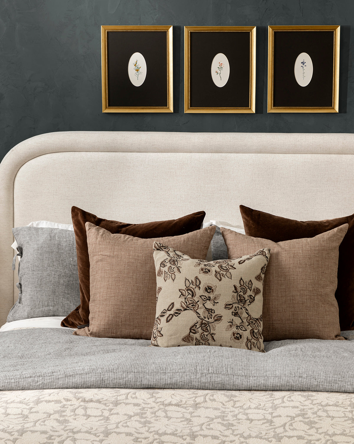 A neatly made bed with gray and beige linens, the Bellevue Velvet Pillow Cover among brown and floral pillows, plus a light upholstered headboard. Three framed botanical prints hang on a dark wall above the bed, by Vendixs
