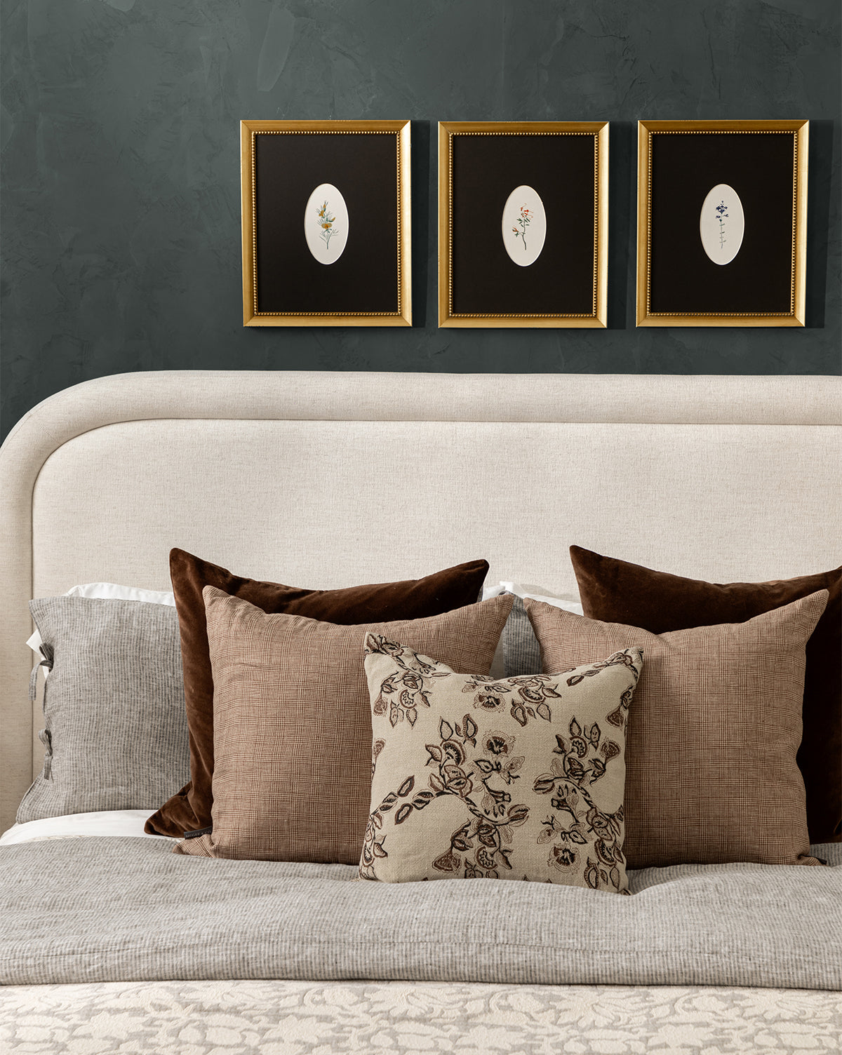A neatly made bed with brown and beige pillows and a beige headboard sits against a dark green wall, topped with three Single Blooms 2 botanical prints in gold-finished wood frames arranged above the bed, by Vendixs