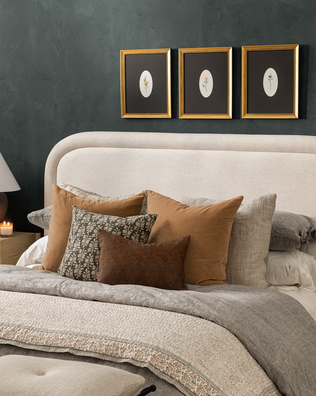 A cozy bedroom with a beige upholstered headboard, layered neutral bedding, assorted throw pillows—including a cotton pillow and the DUPE Bellevue Velvet Pillow Cover—and three botanical prints on a dark green wall above the bed, plus a lamp and candle on the bedside table, by Vendixs