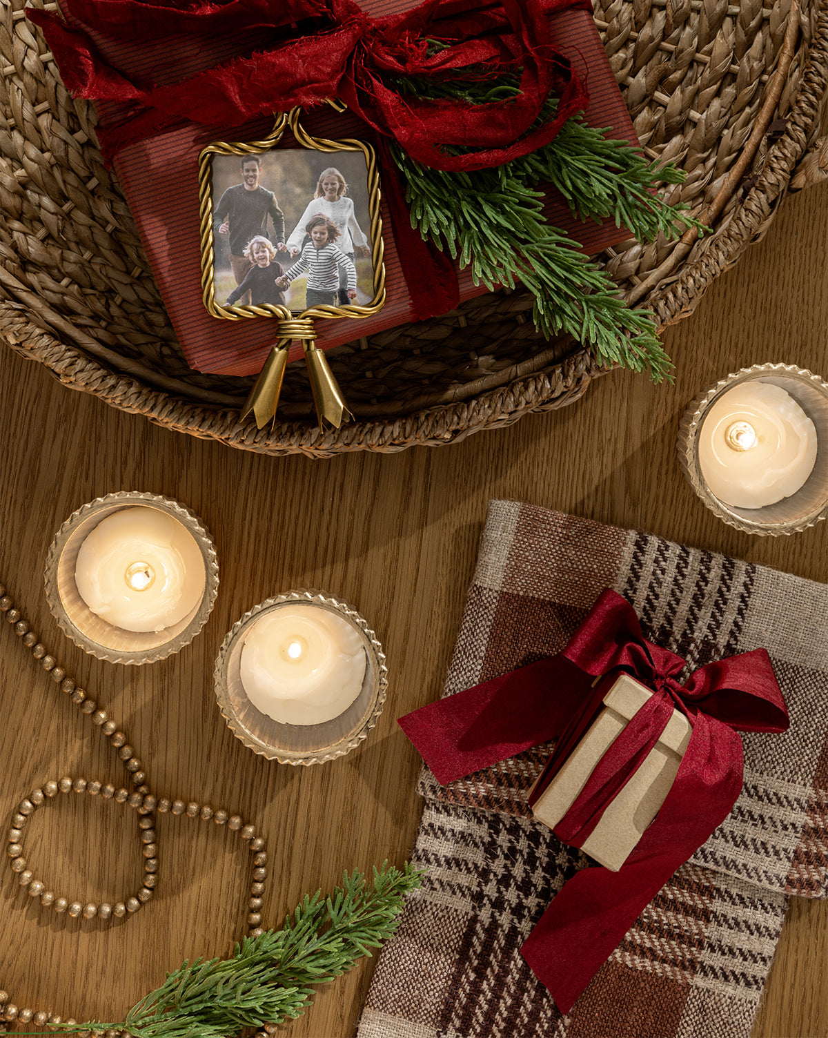 A festive scene with candles, a wrapped gift box, evergreen sprigs, plaid fabric, wooden beads, a framed family photo, and the Tassel Picture Frame Ornament displayed on a wooden table, by Vendixs