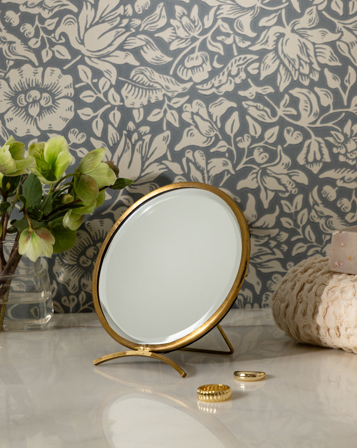 The Antique Brass Folding Mirror stands on a marble countertop beside a potted plant, a textured towel, gold jewelry, and pink soap, all set against floral-patterned wallpaper, by Vendixs