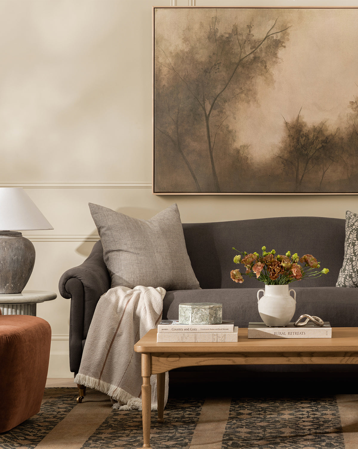 A cozy living room features a dark sofa with gray pillows, a light throw blanket, and a wooden coffee table topped with flowers, books, and a decorative box. Above the sofa hangs Restored by Brittany Schulz on museum-quality canvas, by Vendixs