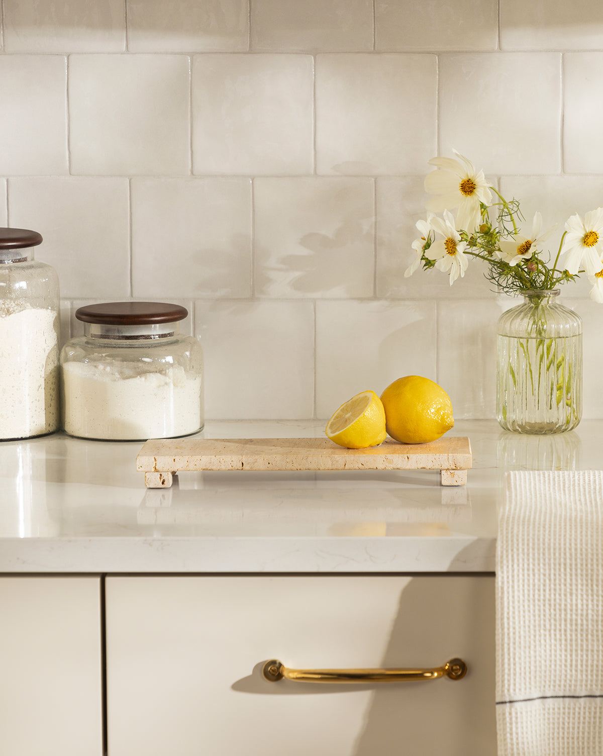 A kitchen counter displays a Beige Travertine Footed Serving Board with two lemons (one halved). Nearby are glass jars of flour, a jar of white flowers, and a hanging towel, all set against a tiled backsplash, by Vendixs