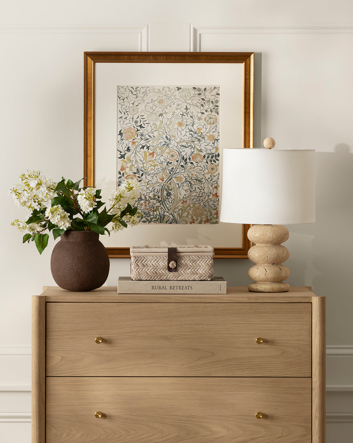 A light wood dresser with antique brass knobs holds a round brown vase of white flowers, stacked books, and the Girault Table Lamp. Above, a framed floral artwork adorns the white paneled wall, enhancing the room’s organic texture, by Vendixs
