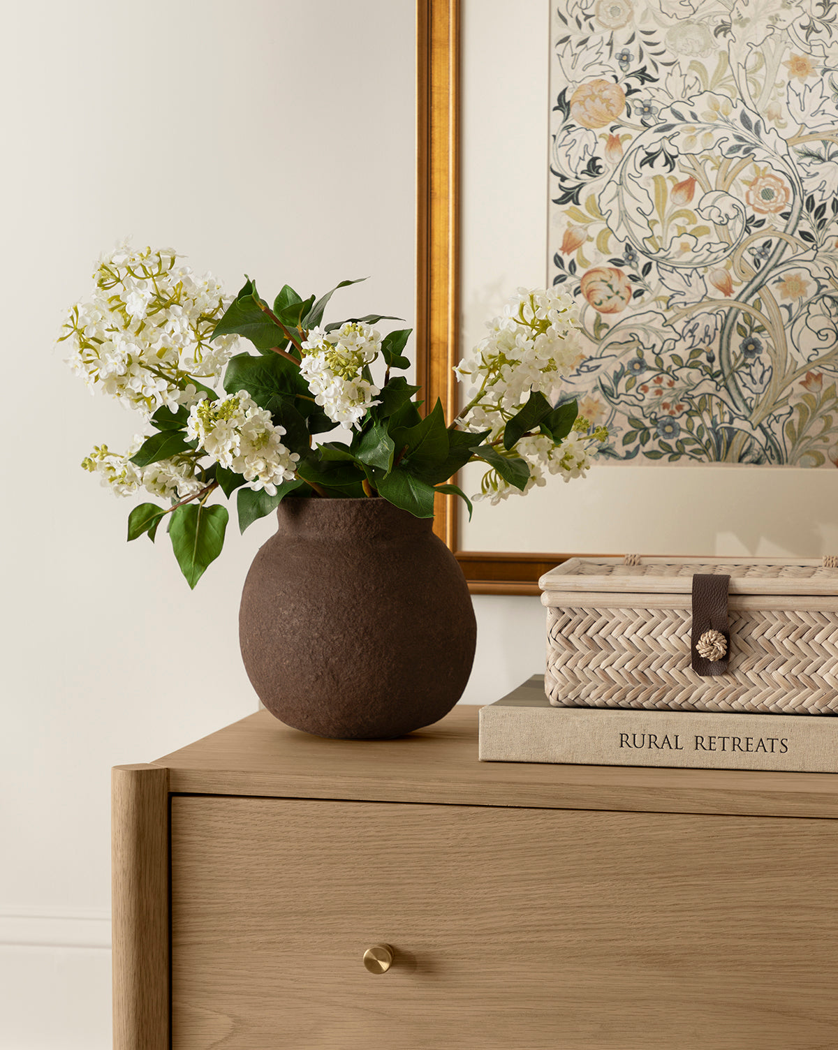 The Frederick Paper Mache Vase with white flowers sits on a light wooden dresser beside a woven box and the book Rural Retreats, while a floral-framed artwork hangs on the wall in the background, by Vendixs