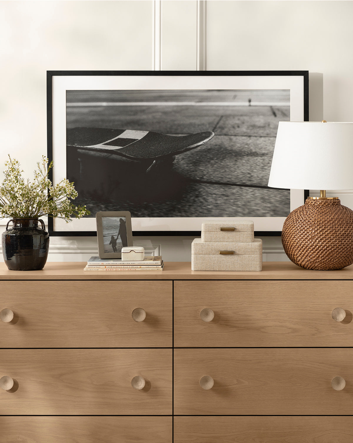 A light wood dresser with round knobs displays the Avedon Table Lamp with a white linen shade, two fabric boxes, a black vase of flowers, a photo frame, and books. Behind them rests a framed black-and-white skateboard photo by Vendixs