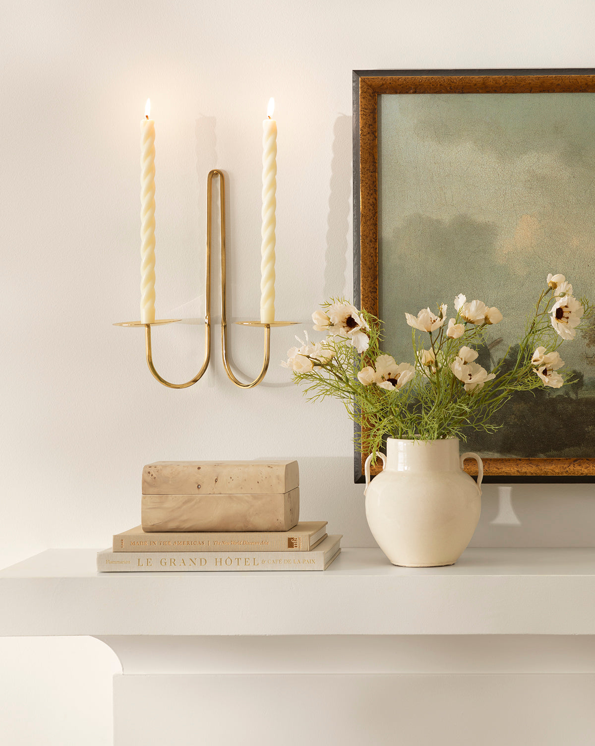 A white mantel with stacked books, a rectangular stone, a cream ceramic pitcher displaying the Cream Cosmos Stem, a framed green landscape painting, and a gold wall candle holder with two lit twisted candles, by Vendixs