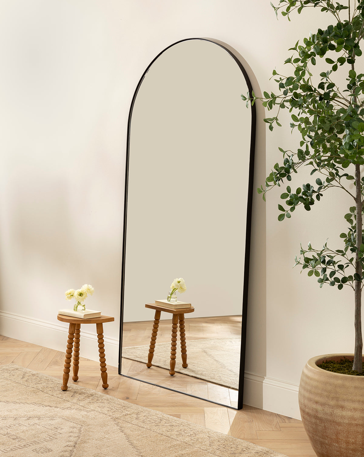 The Gala Floor Mirror, featuring a large arched silhouette and black frame, leans against a light wall. A small turned-leg table with a vase of white flowers and a nearby potted plant complete the look on the wooden floor by Vendixs