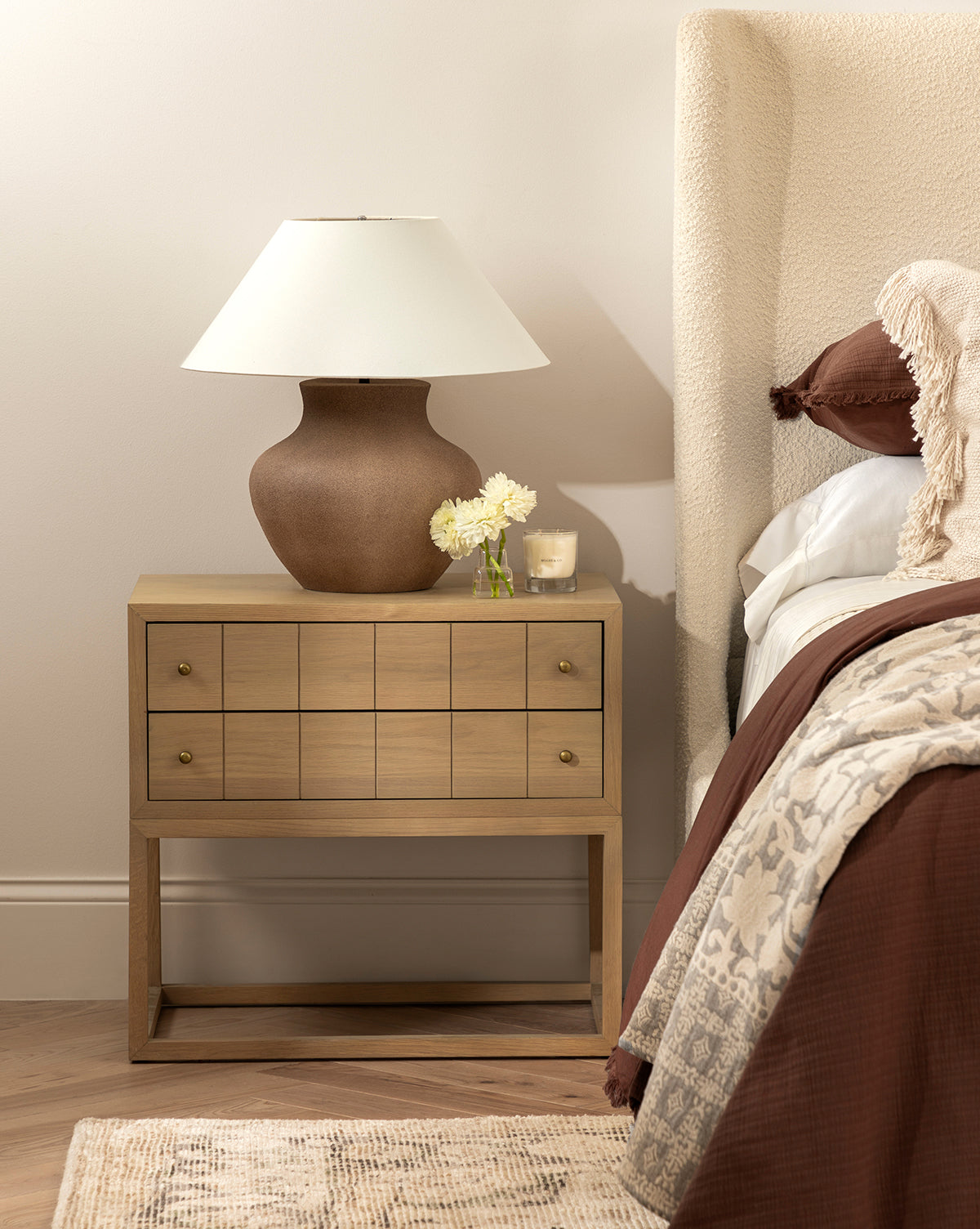 A wooden bedside table holds the Parma Table Lamp, featuring an ivory linen shade and textured ceramic base, next to a glass with white flowers and a candle. It stands beside a bed with brown bedding, a textured headboard, and part of a rug on the wood floor by Vendixs