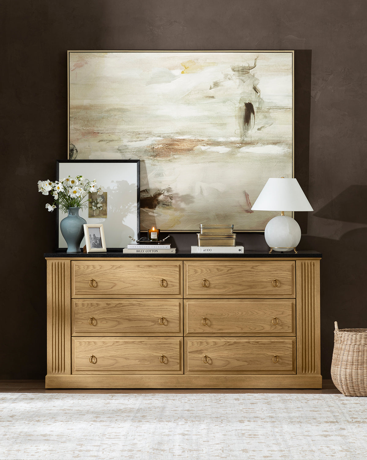 A wooden dresser with six drawers displays the Price Table Lamp with a linen shade, a vase of white flowers, books, candles, and framed art. Above hangs a large abstract painting on a dark wall; a woven basket sits nearby on the floor, by Vendixs