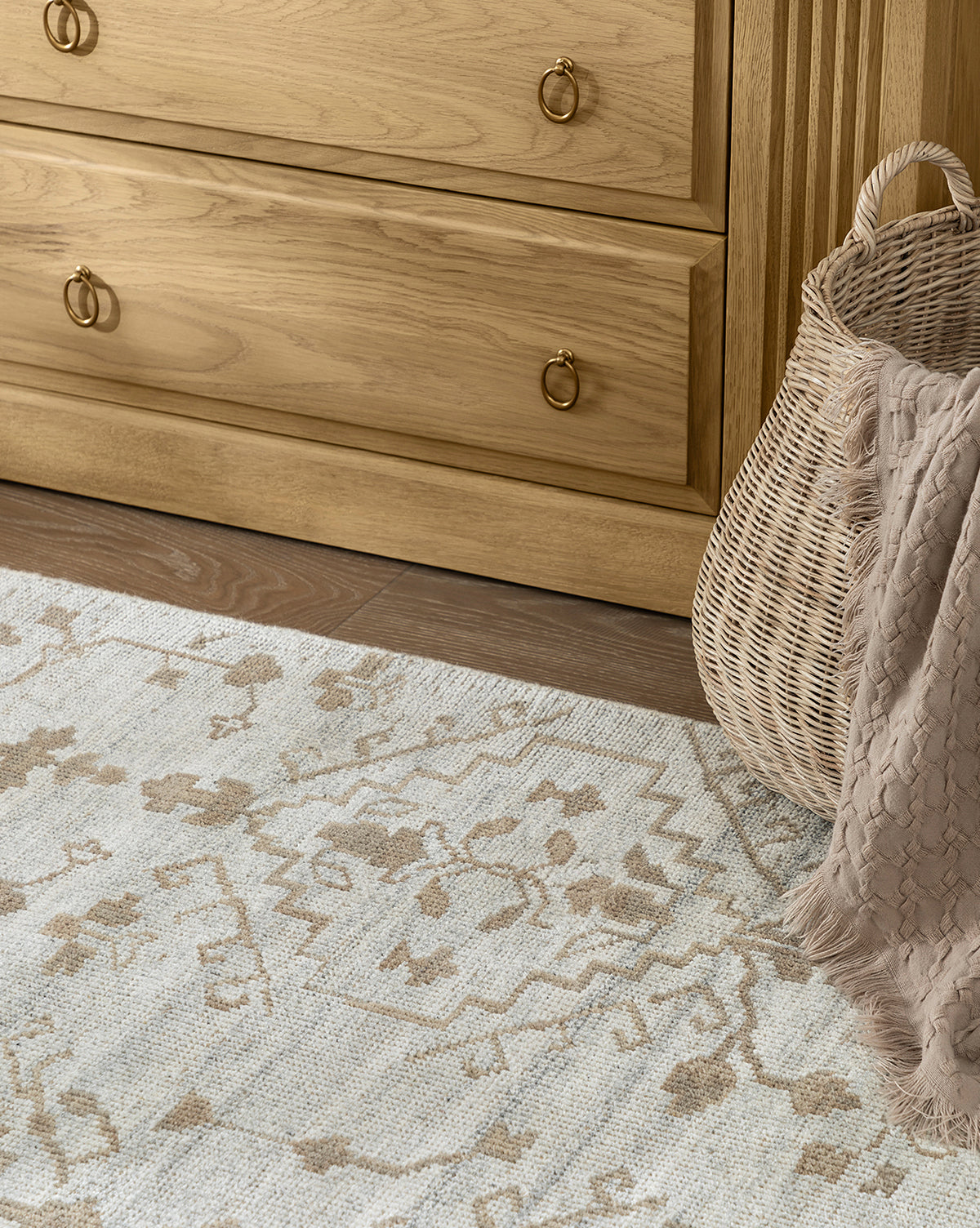 A close-up of the Lexington Hand-Knotted Wool Rug in a light pattern on a wood floor, next to a woven basket with a beige blanket, and in front of a light wood dresser featuring brass handles, by Vendixs