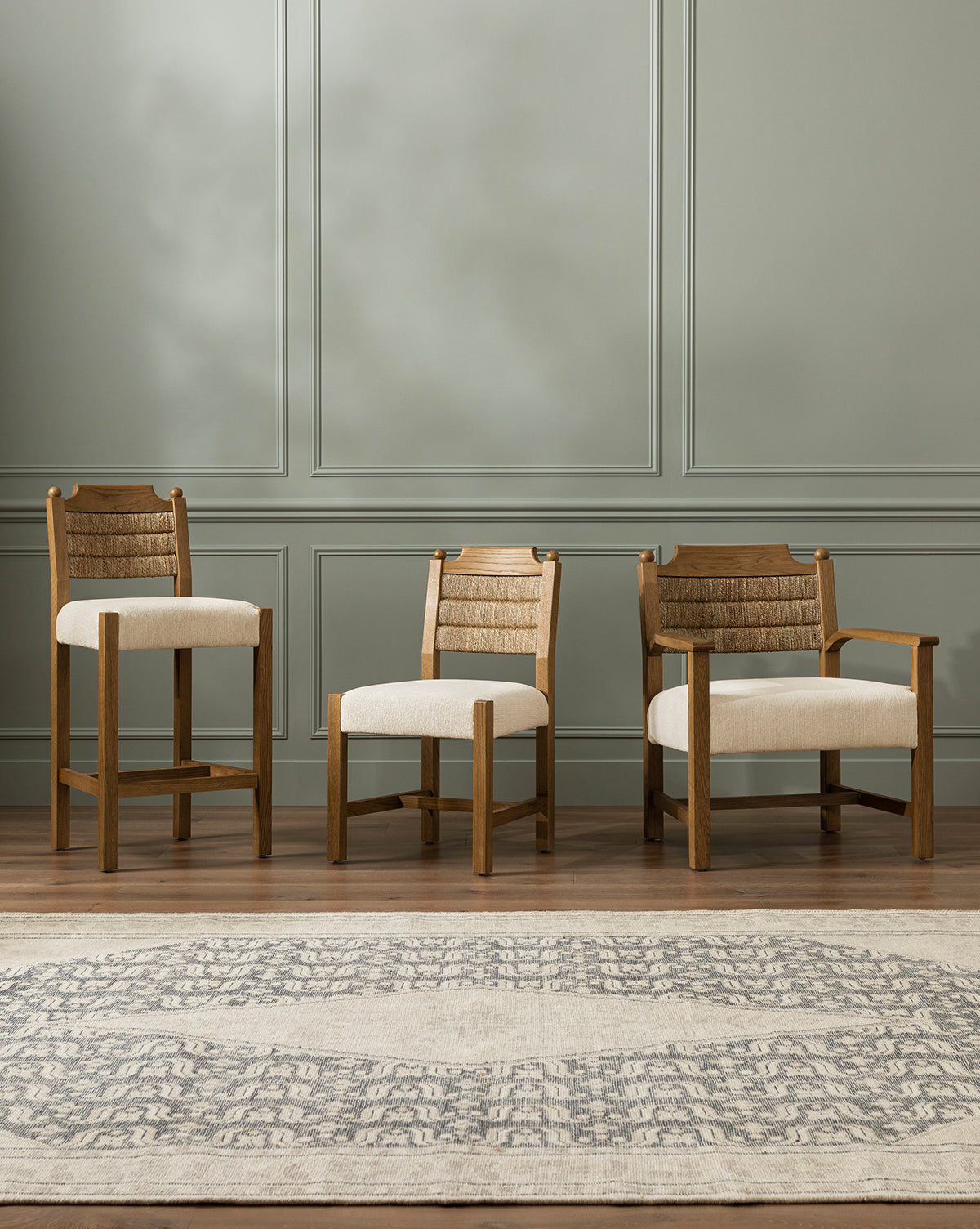 Three Makerspalm Alberta Counter Stools, featuring honey oak wood, beige cushions, and woven rattan backs, are arranged side by side in a room with light green paneled walls and a patterned rug on a wood floor. Vendixs