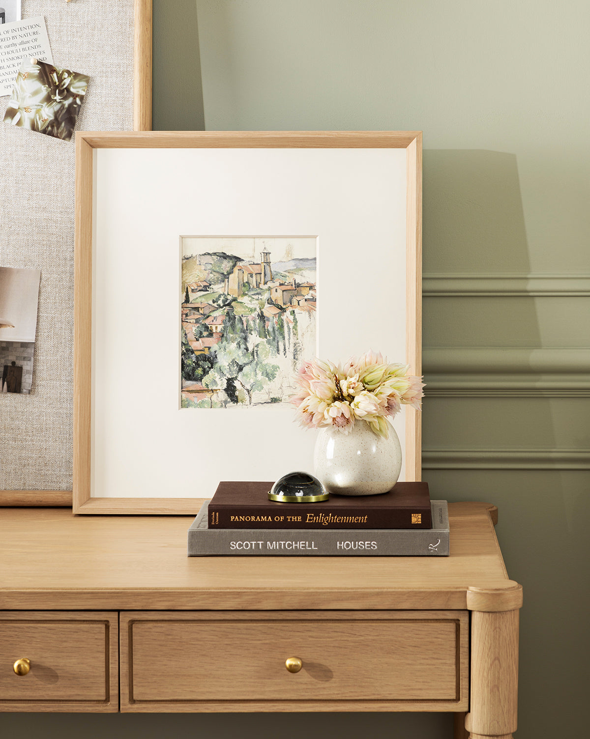 A wooden desk with two drawers displays stacked books, an Erma Bud Vase with pale flowers, and a framed city painting. A bulletin board and sage green wall enhance the inviting home decor in the background, by Vendixs