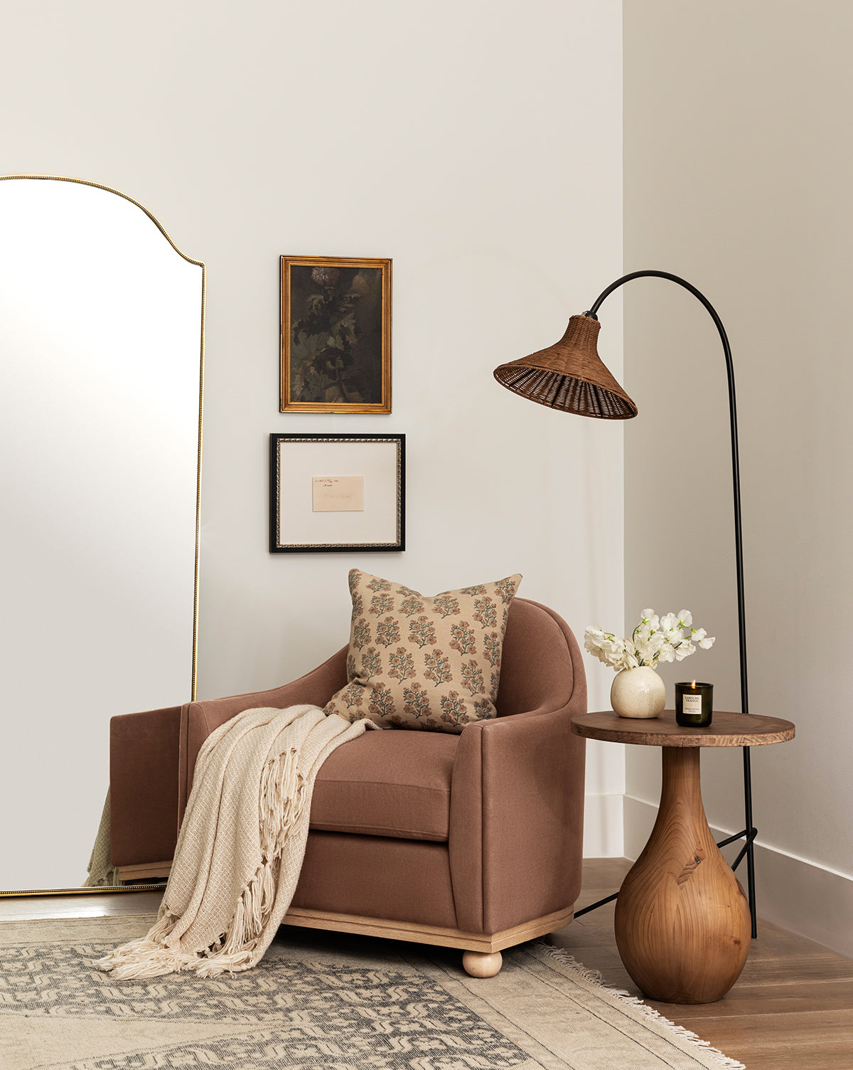 A cozy reading nook with a brown Bunnell Lounge Chair (Ready to Ship), patterned pillow, and cream knit throw beside a round wooden side table topped with flowers and a candle. A floor lamp arches above and framed art decorates the white wall, by Vendixs