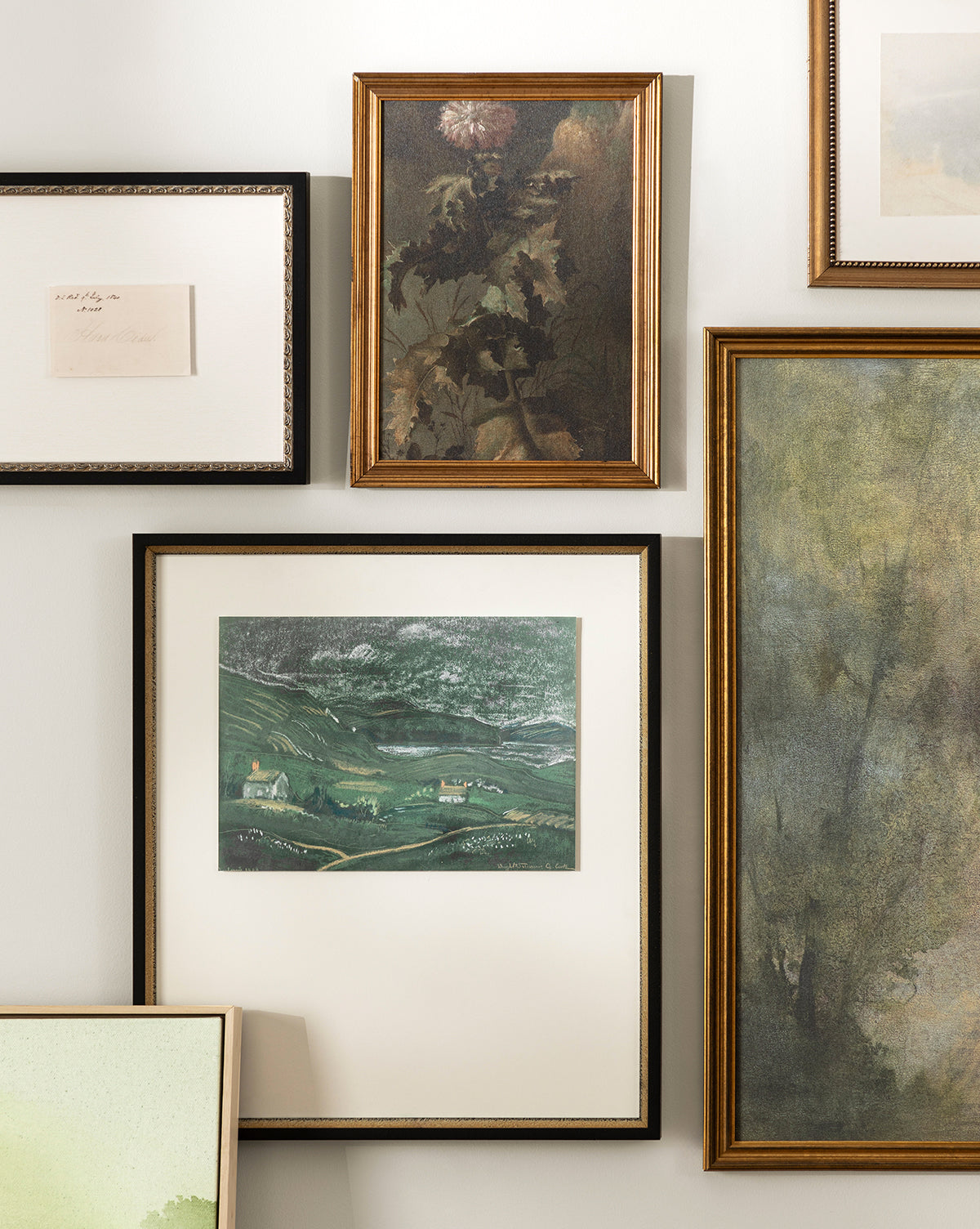 A gallery wall featuring Country Dusk, a giclee print of green hills and houses, paired with a floral painting, an abstract piece, and a framed handwritten note—all closely arranged against a light-colored wall by Vendixs