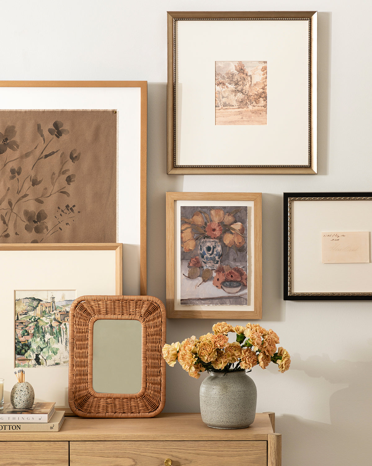 A wooden dresser with a vase of dried flowers and the small Jera Shelf Mirror sits below a gallery wall displaying framed landscape and floral artworks, by Vendixs