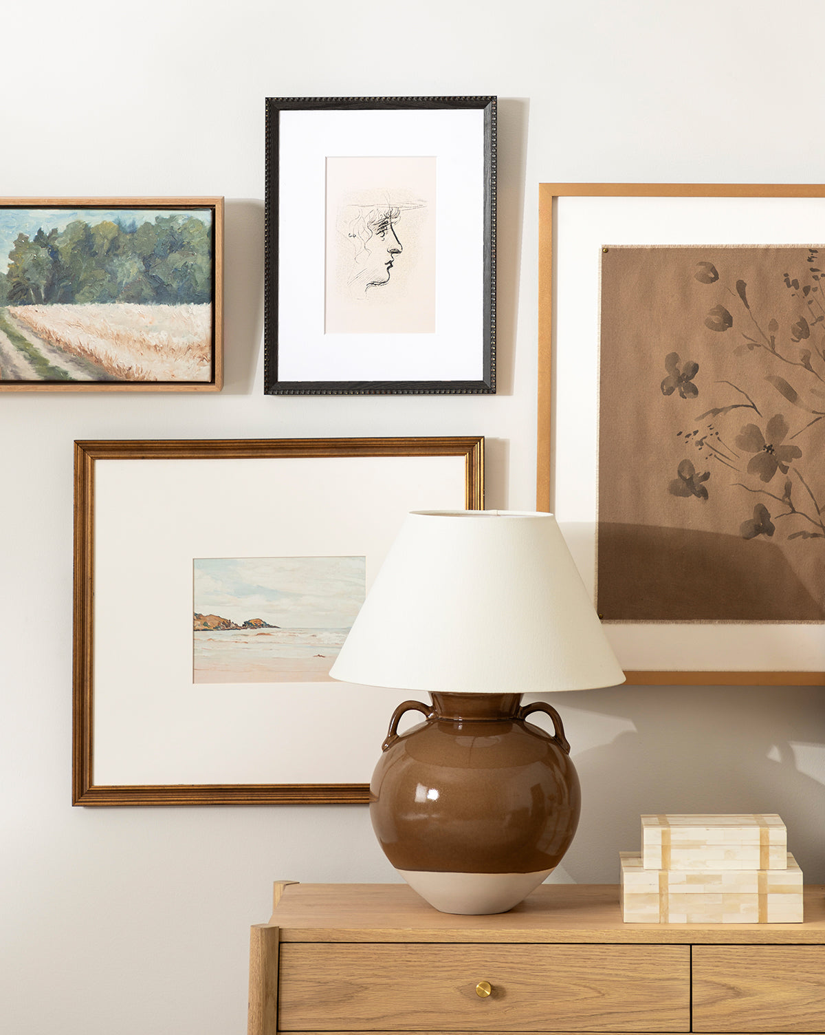 A brown ceramic table lamp with a white shade sits on a wooden dresser, in front of a gallery wall displaying framed landscapes, floral prints, and the Head Study artwork, by Vendixs
