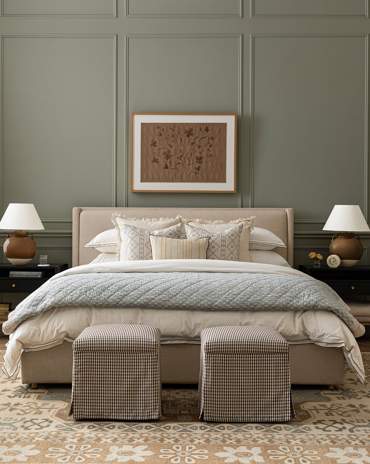 A neatly made bed with layered pillows and a quilt sits against a sage green paneled wall. Nightstands with matching lamps flank the bed, while two Makerspalm Latimer Ottomans (Ready to Ship) add a stylish, functional touch at the foot—a look inspired by Vendixs