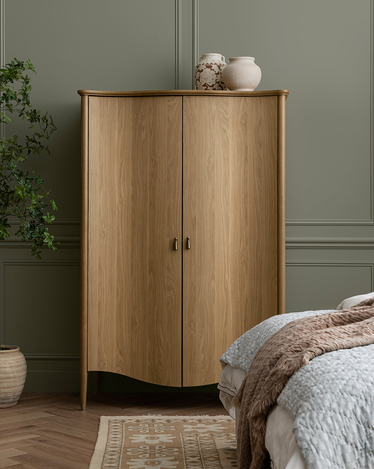 A light wood cabinet with two doors stands against a sage green paneled wall. On top sits the Nola Paper Mache Vase and another decorative piece. A plant, bed with textured blankets, and a patterned rug are partially visible in the room, by Vendixs