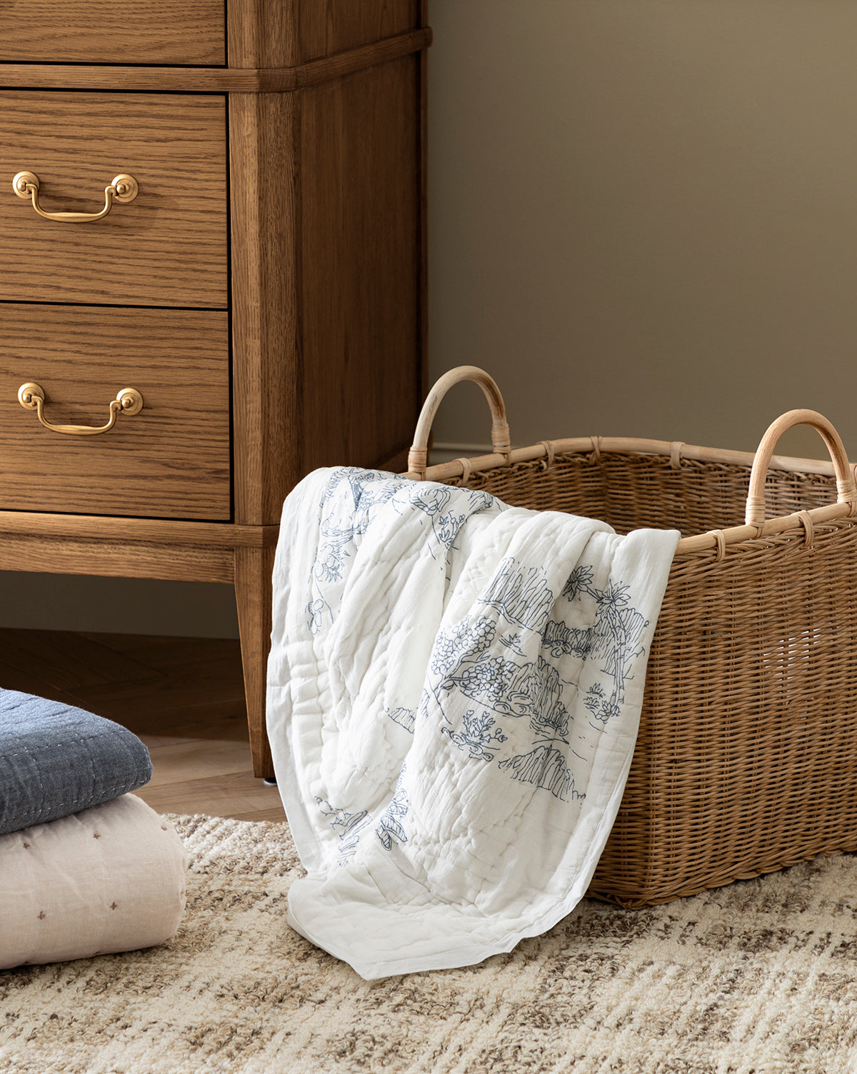 A wicker basket with a Solano Block Print Kids Quilt by Indaba draped over the side sits on a textured rug next to a wooden dresser with brass handles. Folded cotton bedding and kids quilts are stacked neatly on the floor nearby. Vendixs