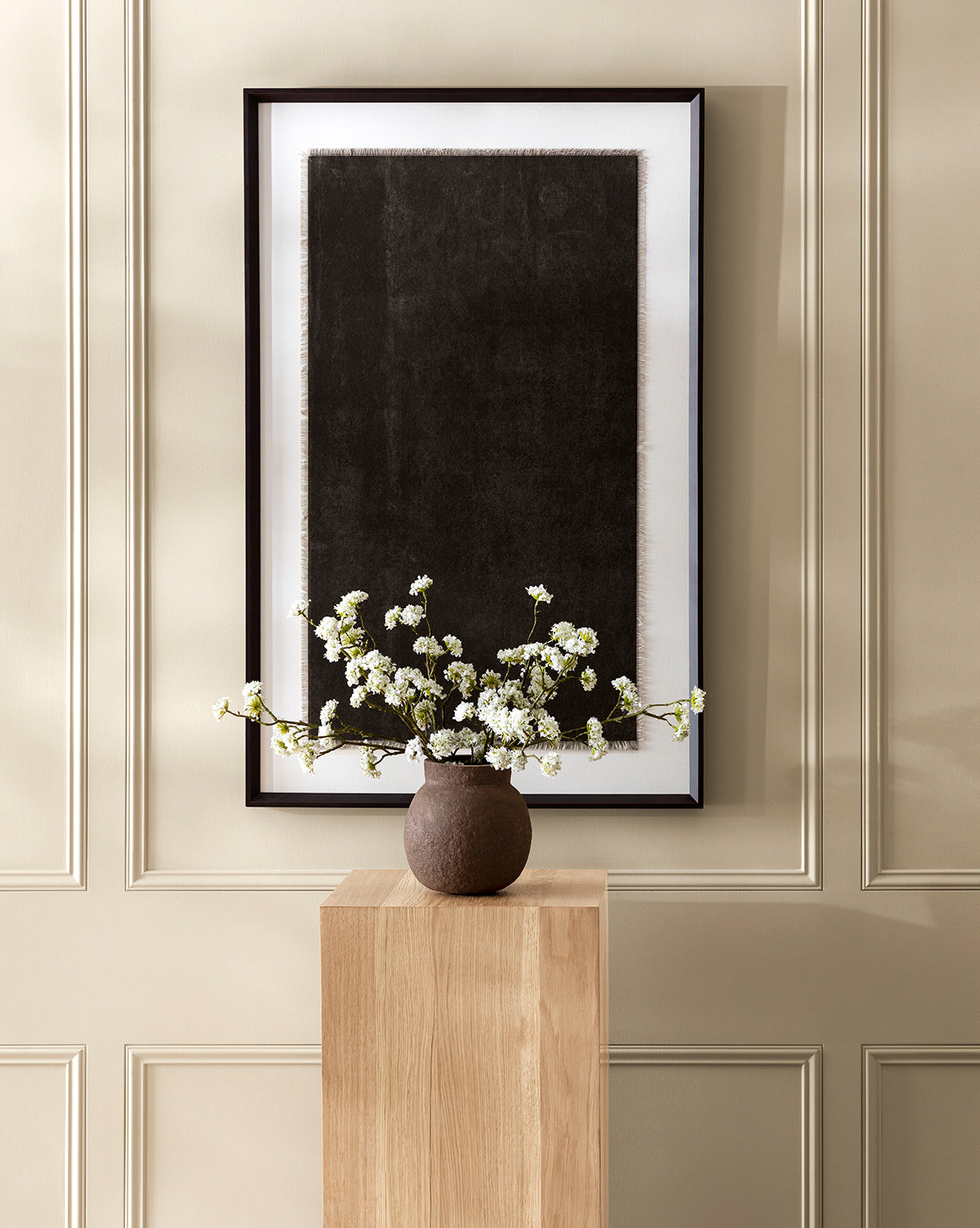 A minimalist interior features the Dark Wash art framed on a beige paneled wall. Below, a wooden pedestal displays a brown vase filled with white flowering branches.