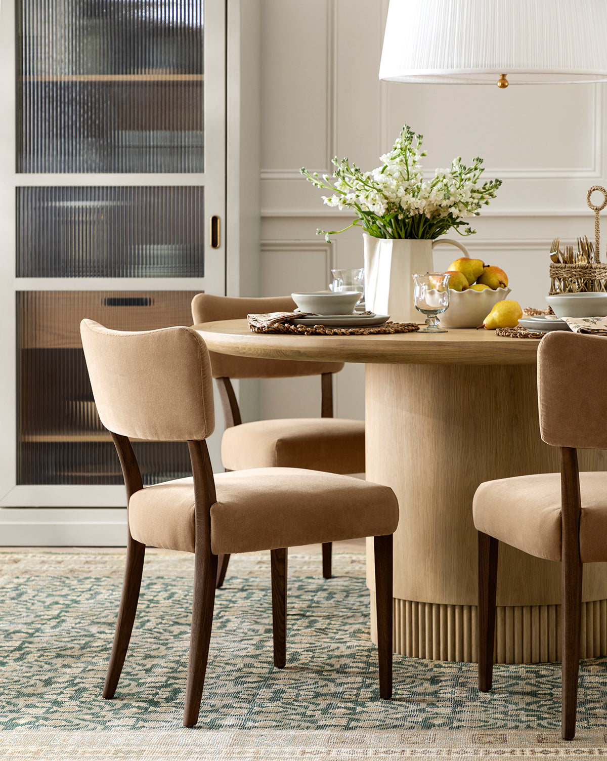 A round wooden dining table with Etienne Dining Chairs (Ready to Ship) upholstered in beige is set with plates, glasses, a vase of white flowers, and a fruit bowl. A glass-fronted cabinet and patterned rug are in the background, by McGee & Co.
