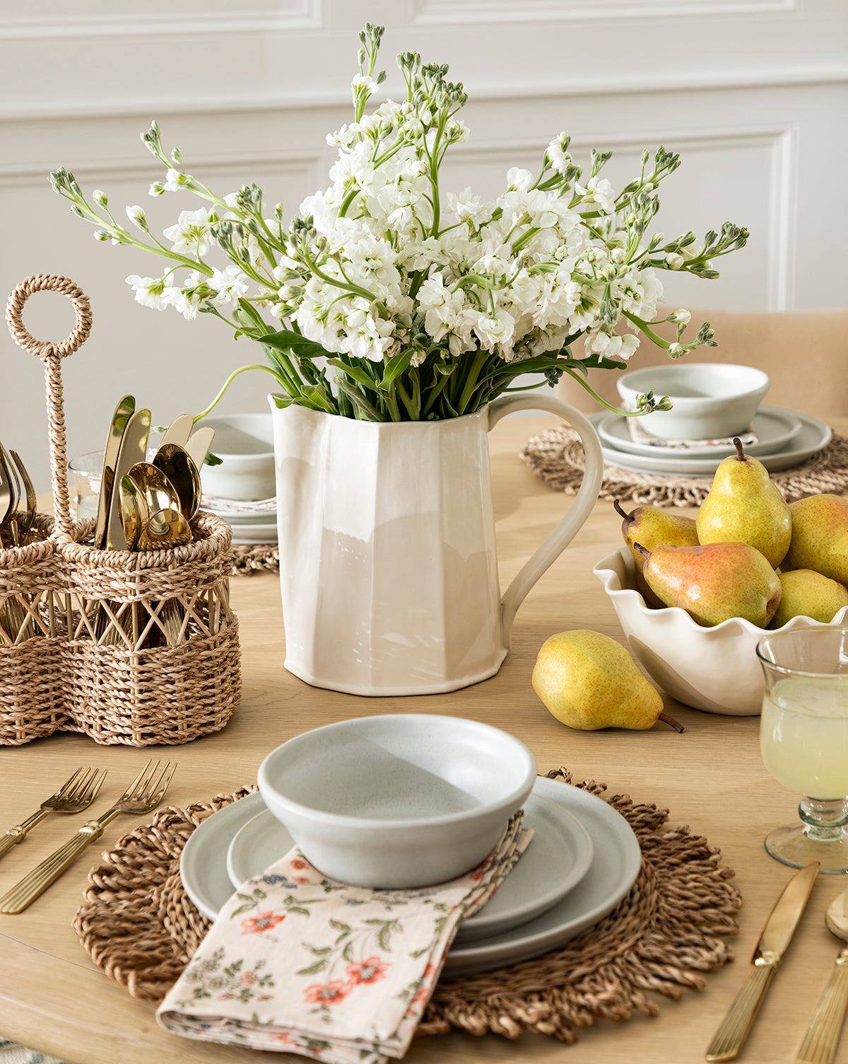 A dining table set for a meal features gray dishes, gold cutlery in a basket, a floral napkin, rattan placemats, the Fielding Ceramic Pitcher with white flowers, fresh pears, and lemonade for cozy rustic kitchen decor, by Vendixs