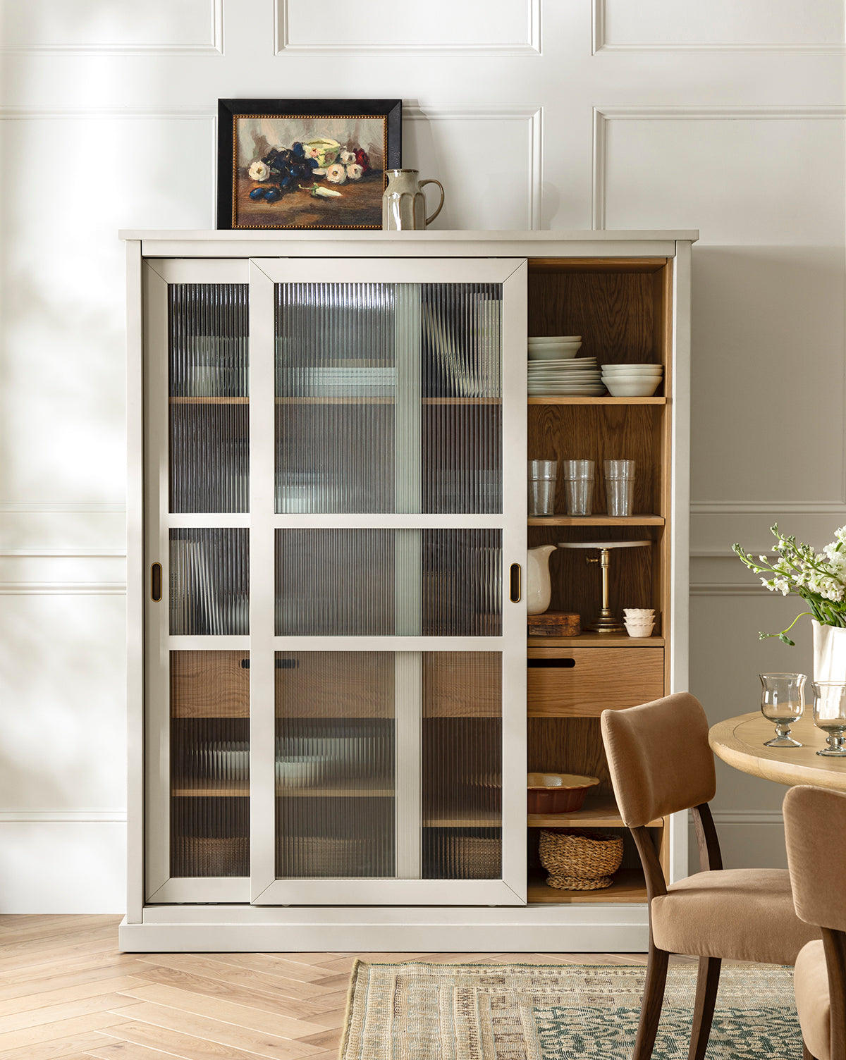 A white cabinet with ribbed glass doors showcases kitchenware, while Tableside by Joyce Washor, plus a jug and cup, adorn the top. A set dining table and chairs are arranged in the foreground, by Vendixs