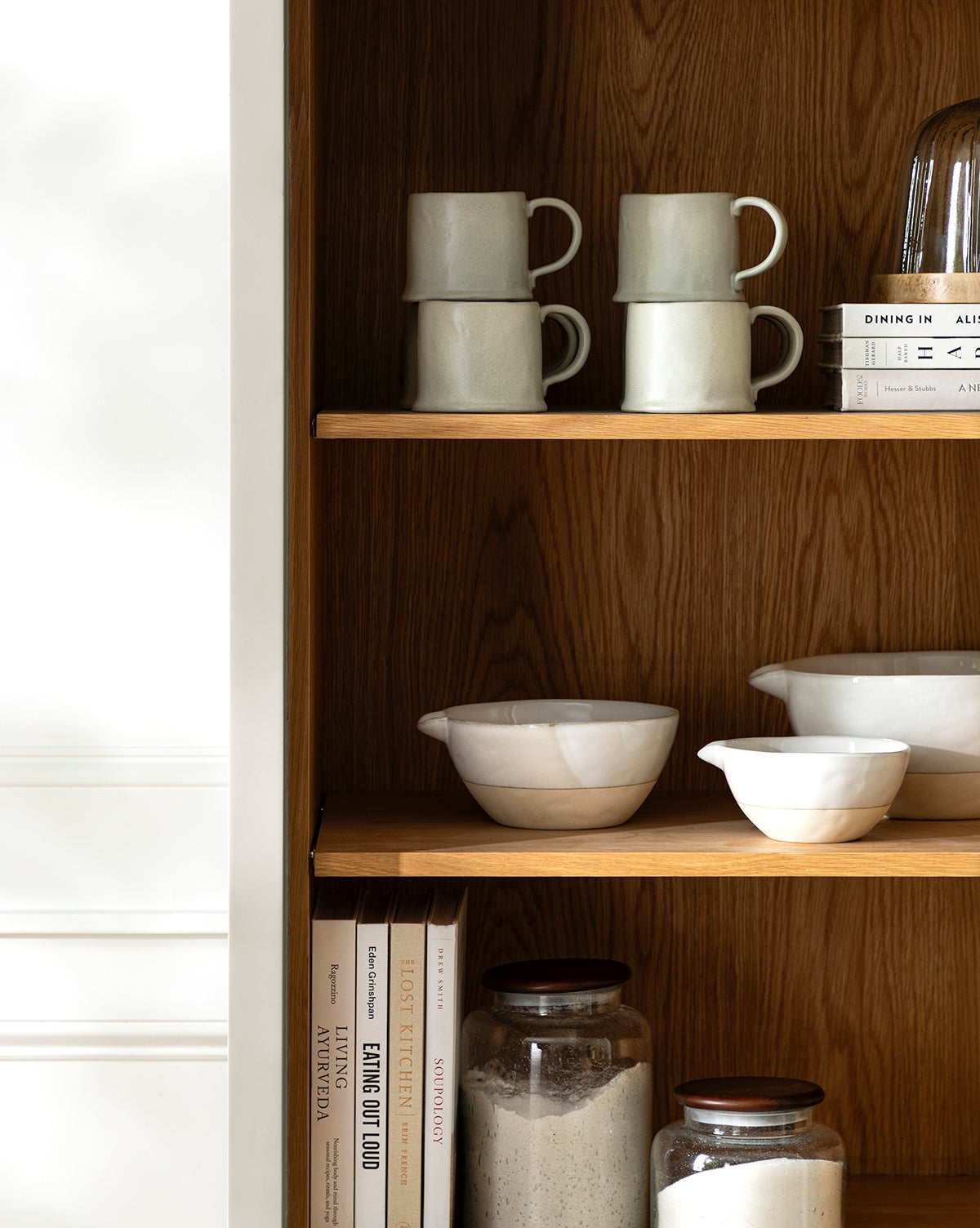 A wooden kitchen shelf displays stacked mugs, white ceramic bowls, vintage baking books, two glass jars with dry ingredients, a set of Karina Stoneware Batter Bowls (Set of 4), and a stack of plates against a warm wood grain background, by Vendixs