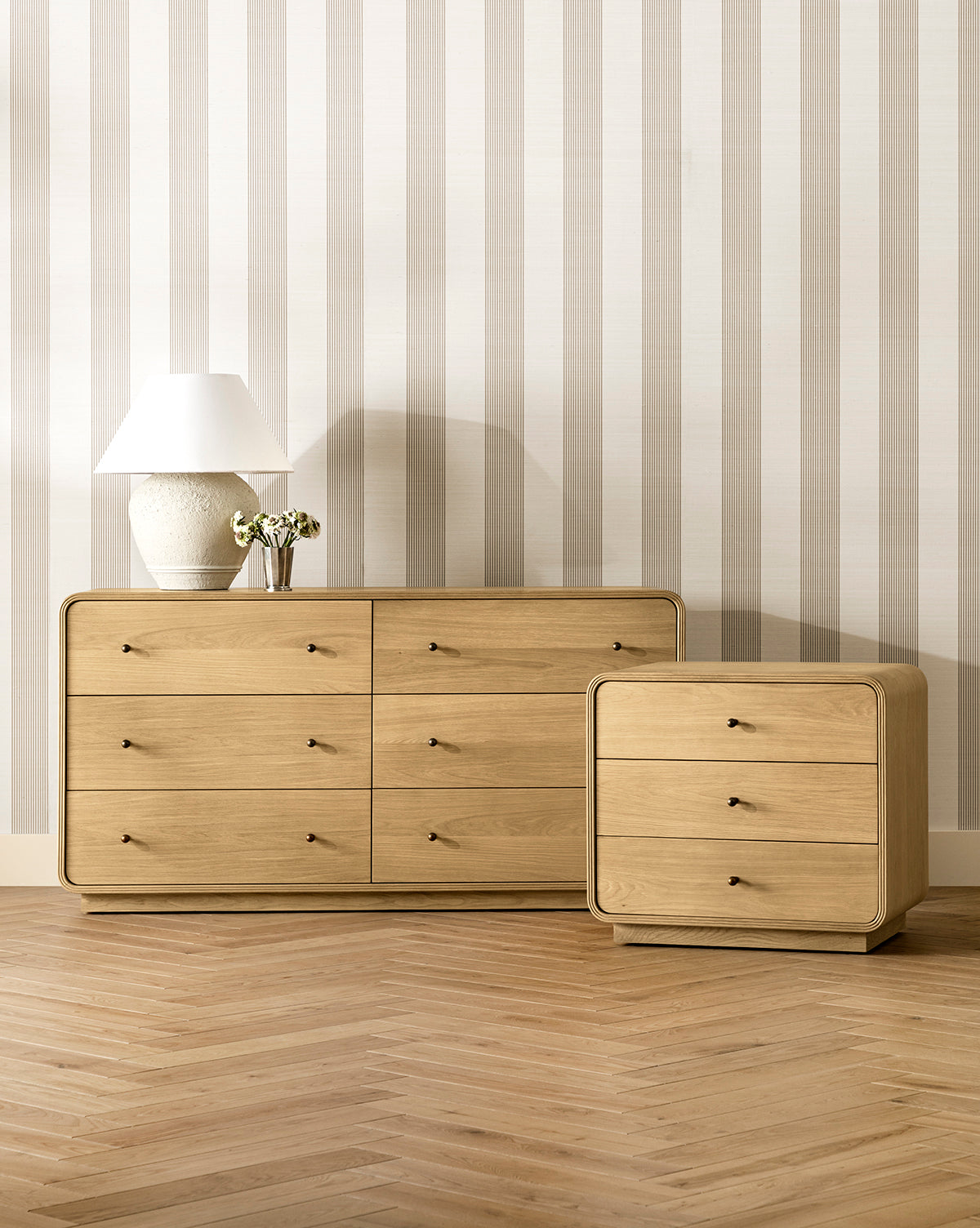 A modern room featuring light wood floors, Leah Striped Sisal Wallpaper by York Wallcoverings, a six-drawer dresser, a matching three-drawer nightstand, a white lamp, and a small vase with flowers by Vendixs