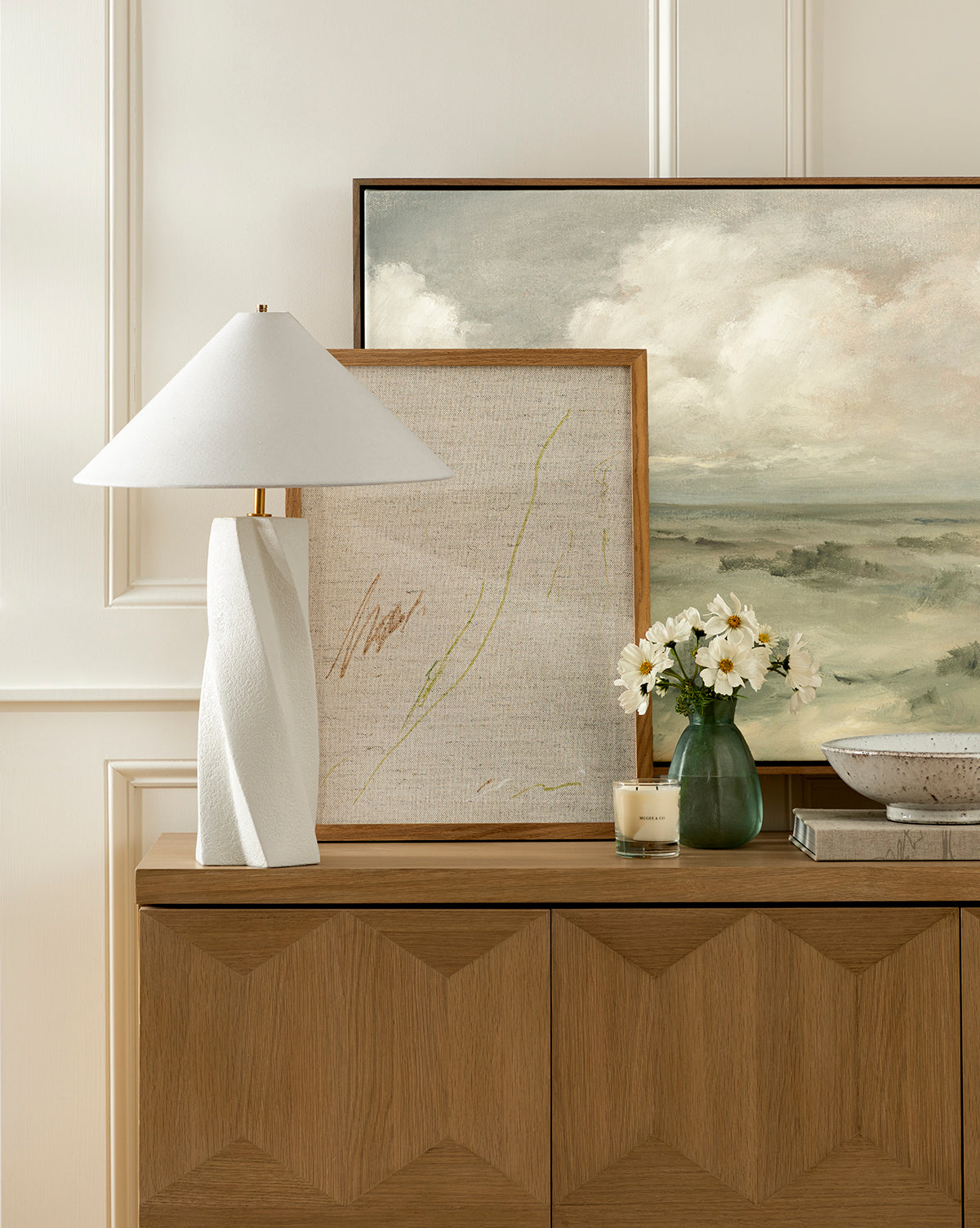 A wooden sideboard displays the modern Bettencourt Table Lamp with a linen shade, two framed art pieces, a small vase of white flowers, a candle, and a decorative bowl against a cream-colored paneled wall, by Vendixs