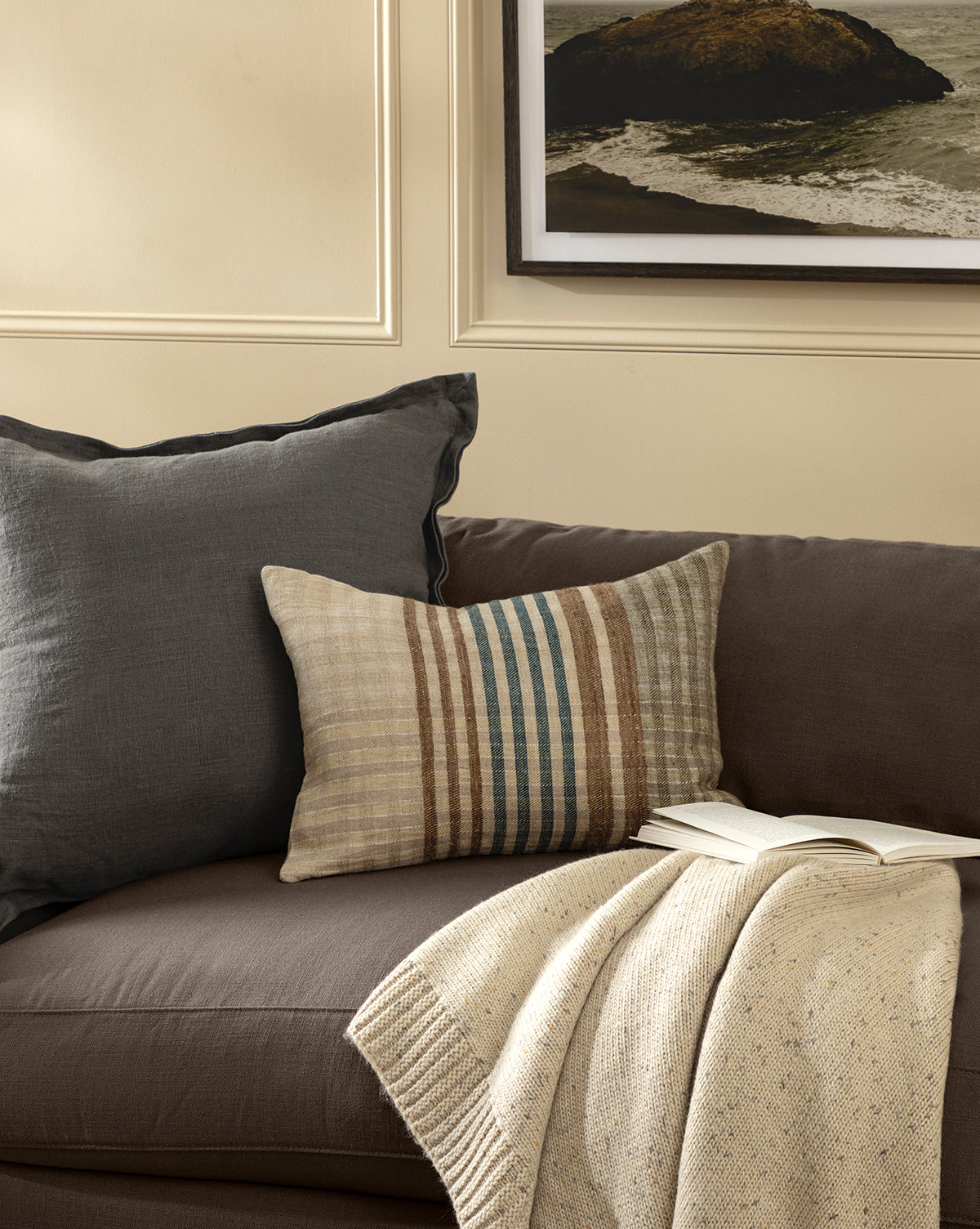 A brown sofa with a gray pillow, a striped beige pillow, the Makerspalm Otis Knit Throw in cream, and an open book. Beige walls and a framed rocky shore print hang above the sofa, blending seamlessly with pieces from Vendixs