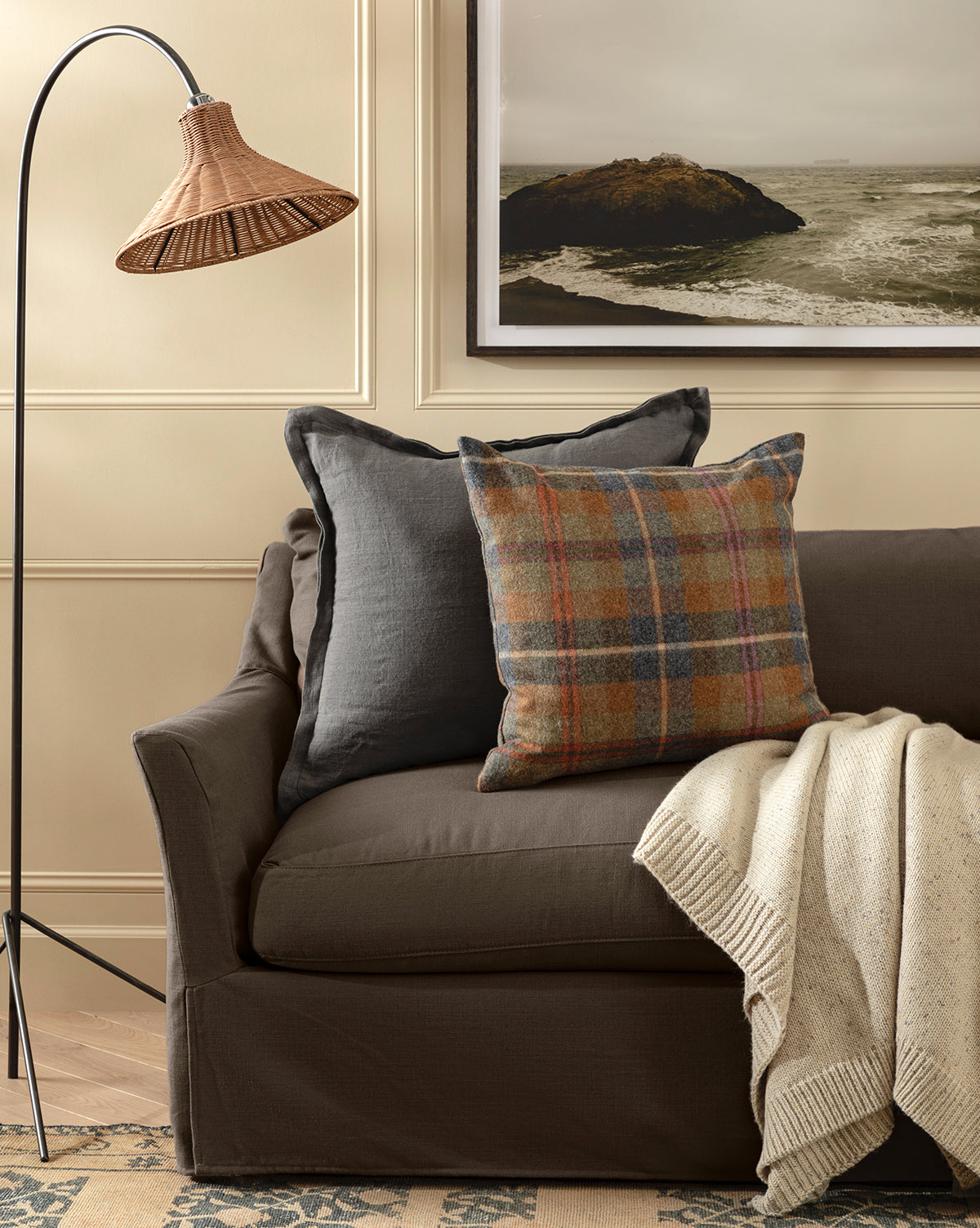 A cozy living room scene with a brown sofa, Liam Double Flange Pillow Cover, plaid pillow, cream knit throw, woven floor lamp, and framed coastal landscape on the wall, by Vendixs