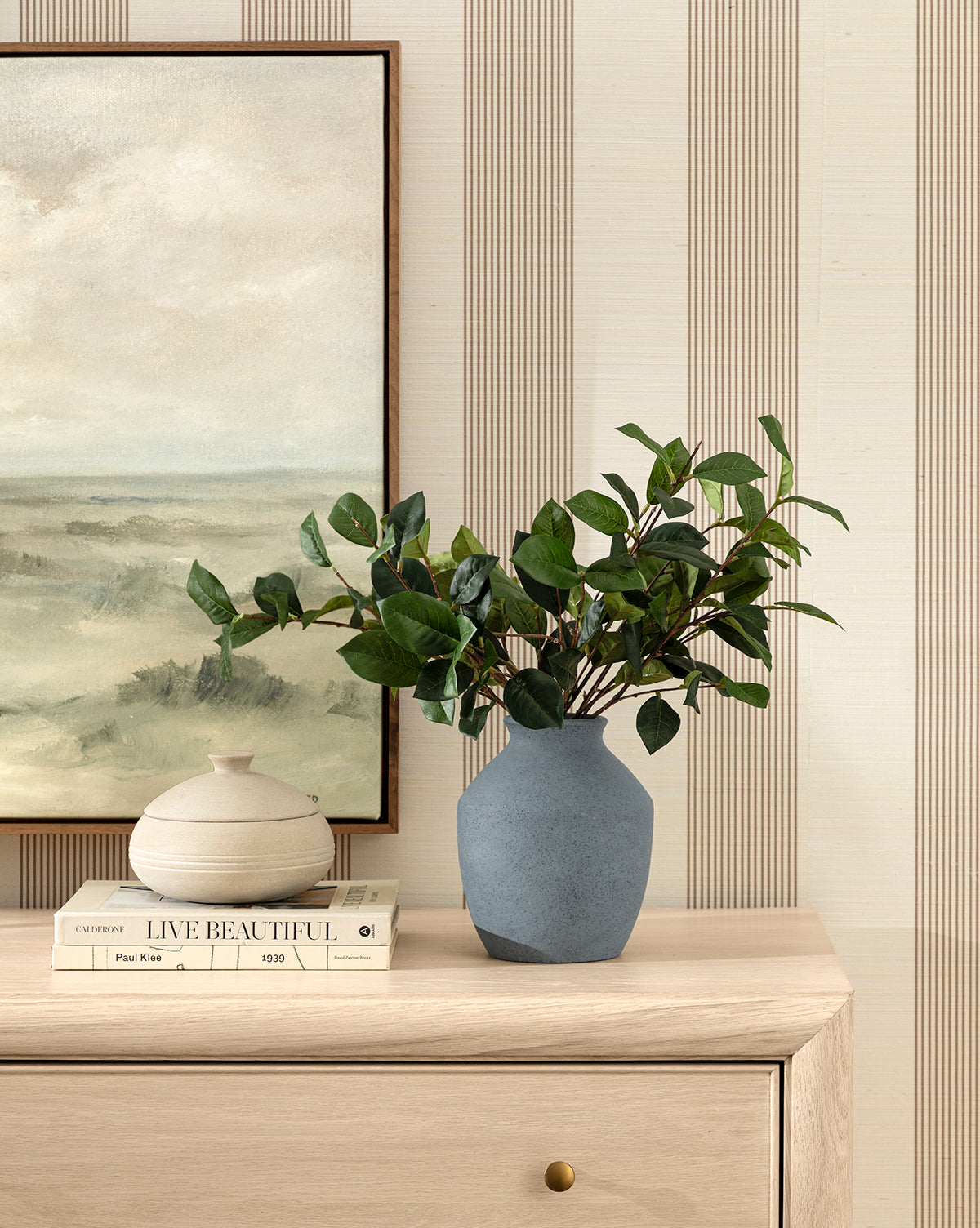 A light wood dresser displays stacked books, a round cream vase, and a blue ceramic vase with a Faux Laurel Leaf Branch. A landscape painting and striped wallpaper complete the serene, modern atmosphere, by Vendixs
