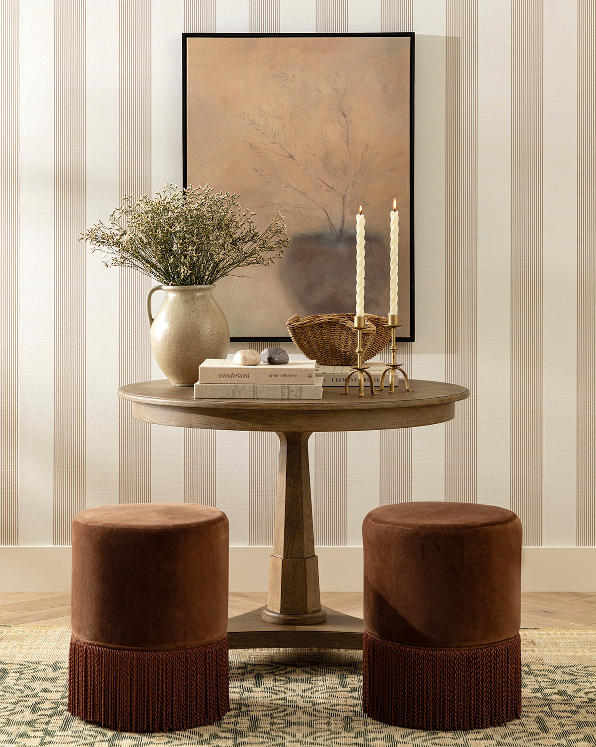 A round wooden table with two brown velvet stools, a vase with greenery, stacked books, a candle holder with lit candles, and a basket sit before neutral artwork and Leah Striped Sisal Wallpaper by York Wallcoverings—finishing the scene with timeless touches from Vendixs