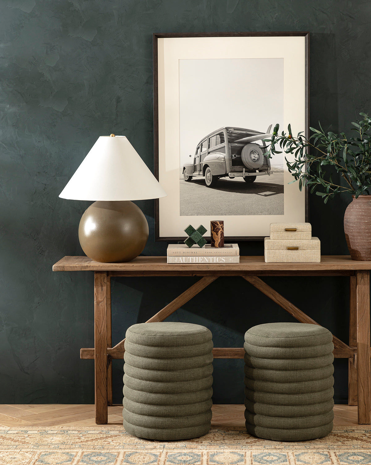 A wooden console table displays the Tess Large Ceramic Table Lamp, books, decorative boxes, and a potted plant. Above hangs a framed black-and-white vintage car photo. Two stacked round green ottomans sit beneath the table, by Vendixs