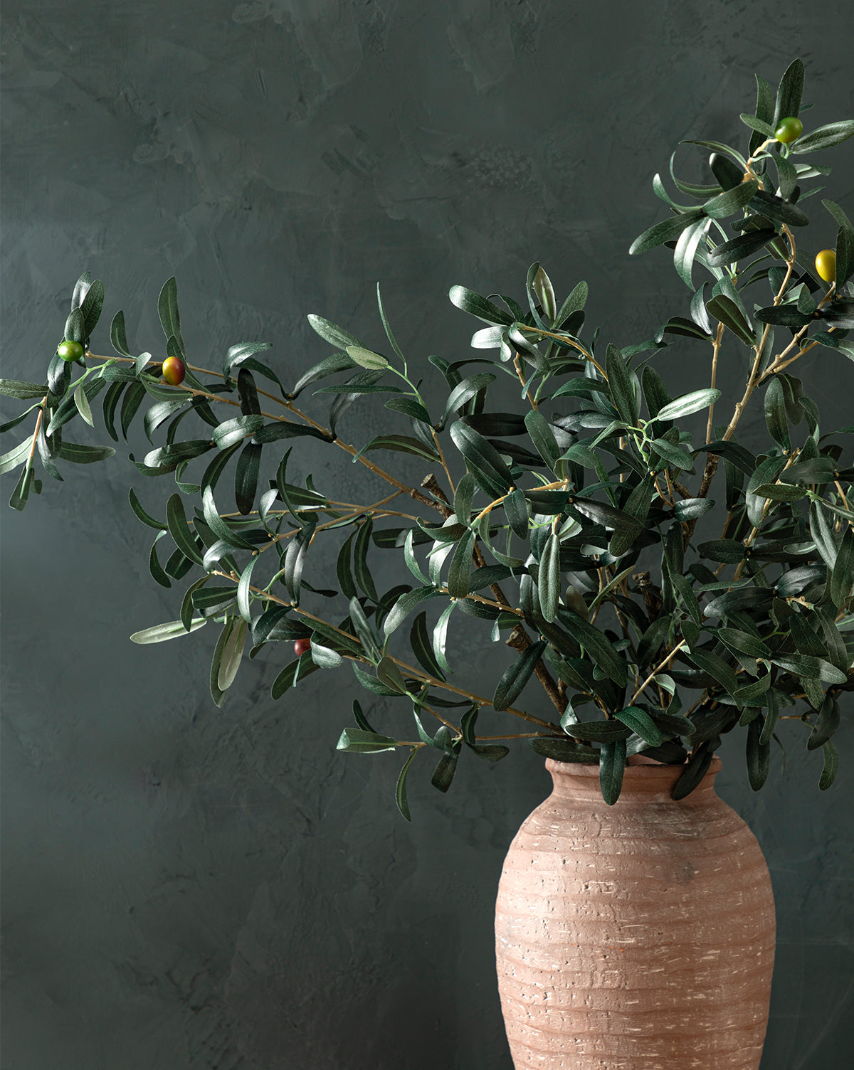 A Faux Olive Branch with dark green leaves and ripening olives is arranged in a terracotta vase, set against a textured dark green background, by Vendixs