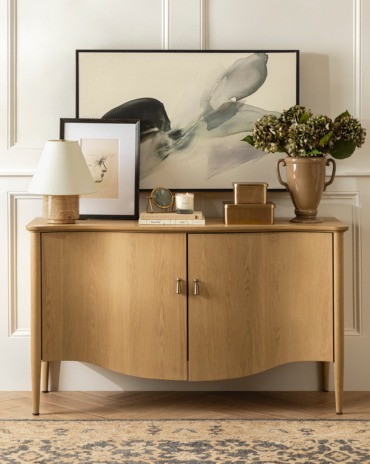 A wooden sideboard stands against a paneled wall, topped with a lamp, gold boxes, candle, clock, vase of green hydrangeas, and framed Head Study artwork featuring expressive linework. A patterned rug covers the floor by Vendixs