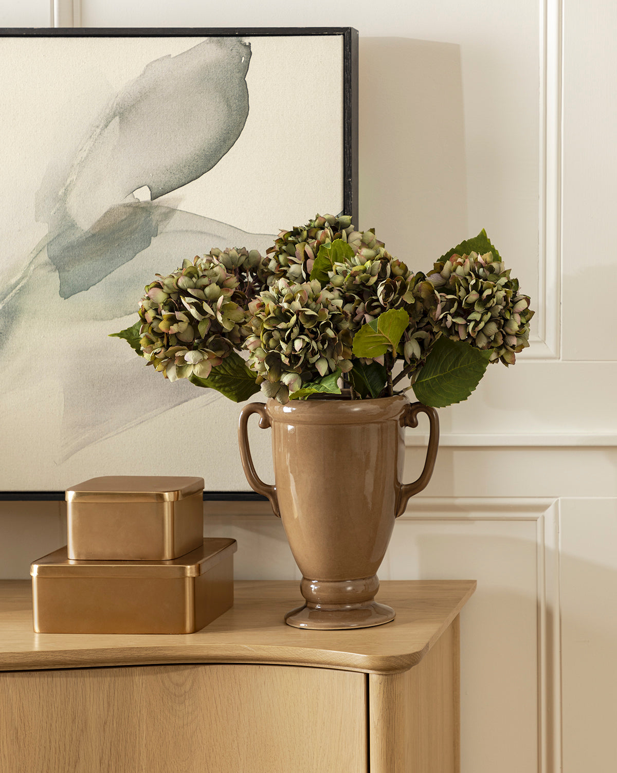 A brown ceramic vase with a Green Hydrangea Stem sits on a light wood table beside two gold rectangular boxes, with a minimalist floral painting in the background, by Vendixs