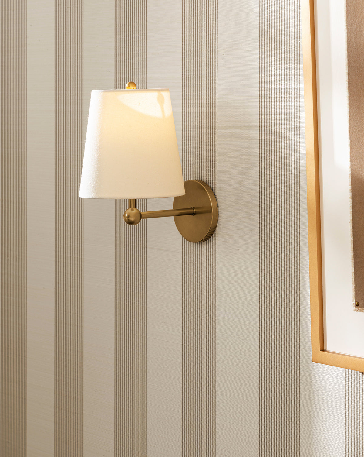 A brass wall-mounted lamp with a white shade glows against Leah Striped Sisal Wallpaper by York Wallcoverings. A gold-framed picture is partially visible on the right side of the image, Vendixs