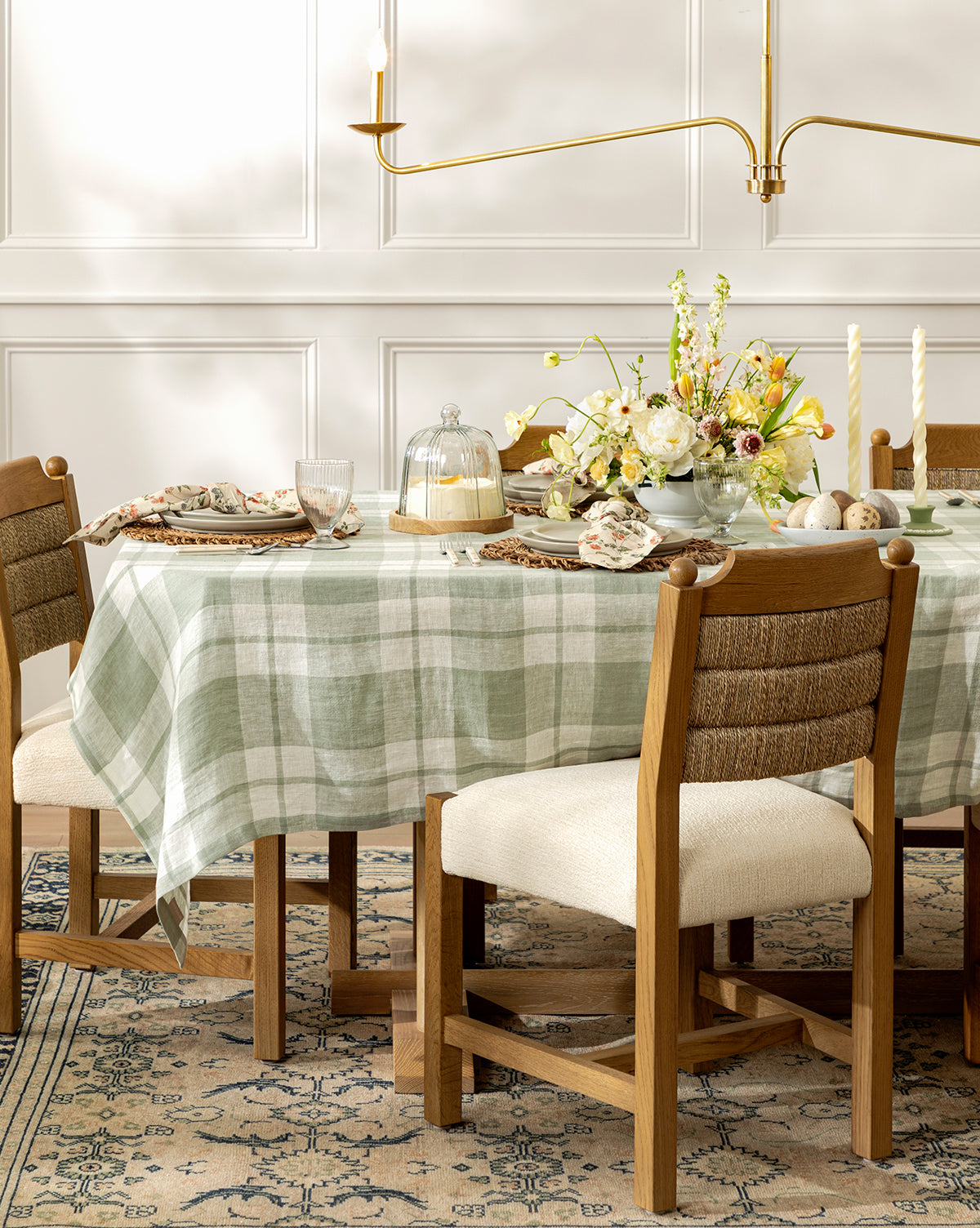 A dining table with a green and white checkered tablecloth is set for spring, surrounded by Alberta Dining Chairs on a patterned rug, with dishes, glasses, candles, flowers, and a cake under a glass dome, by Vendixs